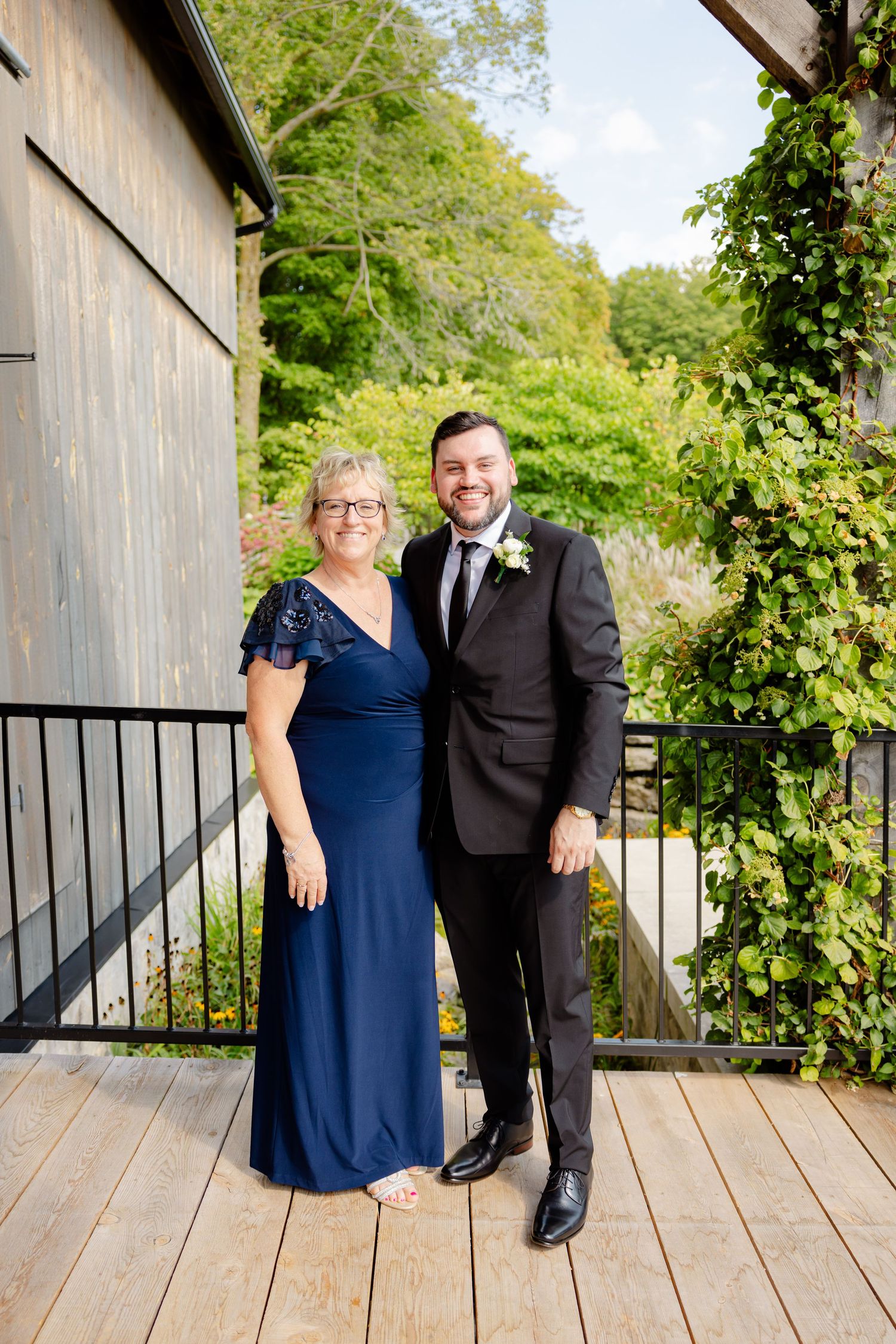 Formal portrait of two people in evening wear on wooden deck with lush garden background.