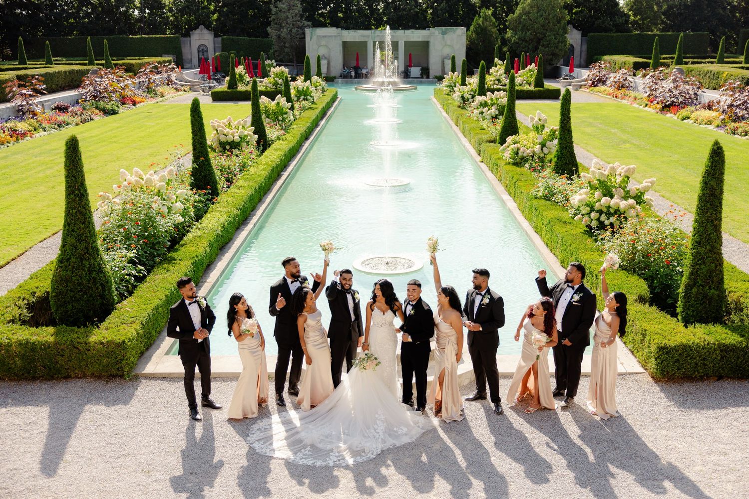 Celebratory wedding party jumping photo in front of a beautiful formal garden water feature.
