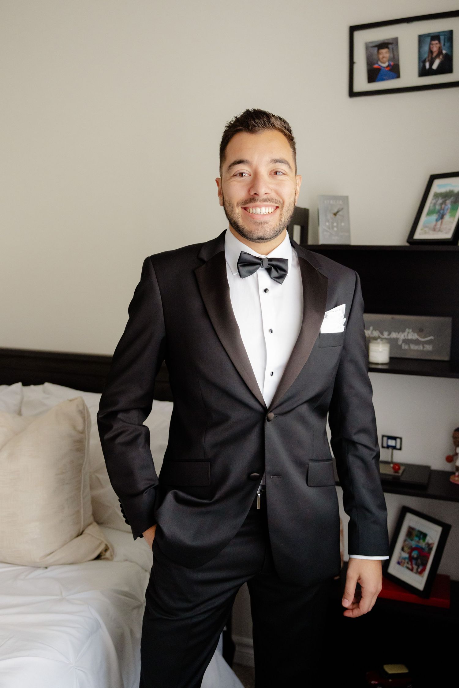 Person in classic black tuxedo and bow tie poses confidently in bedroom setting.
