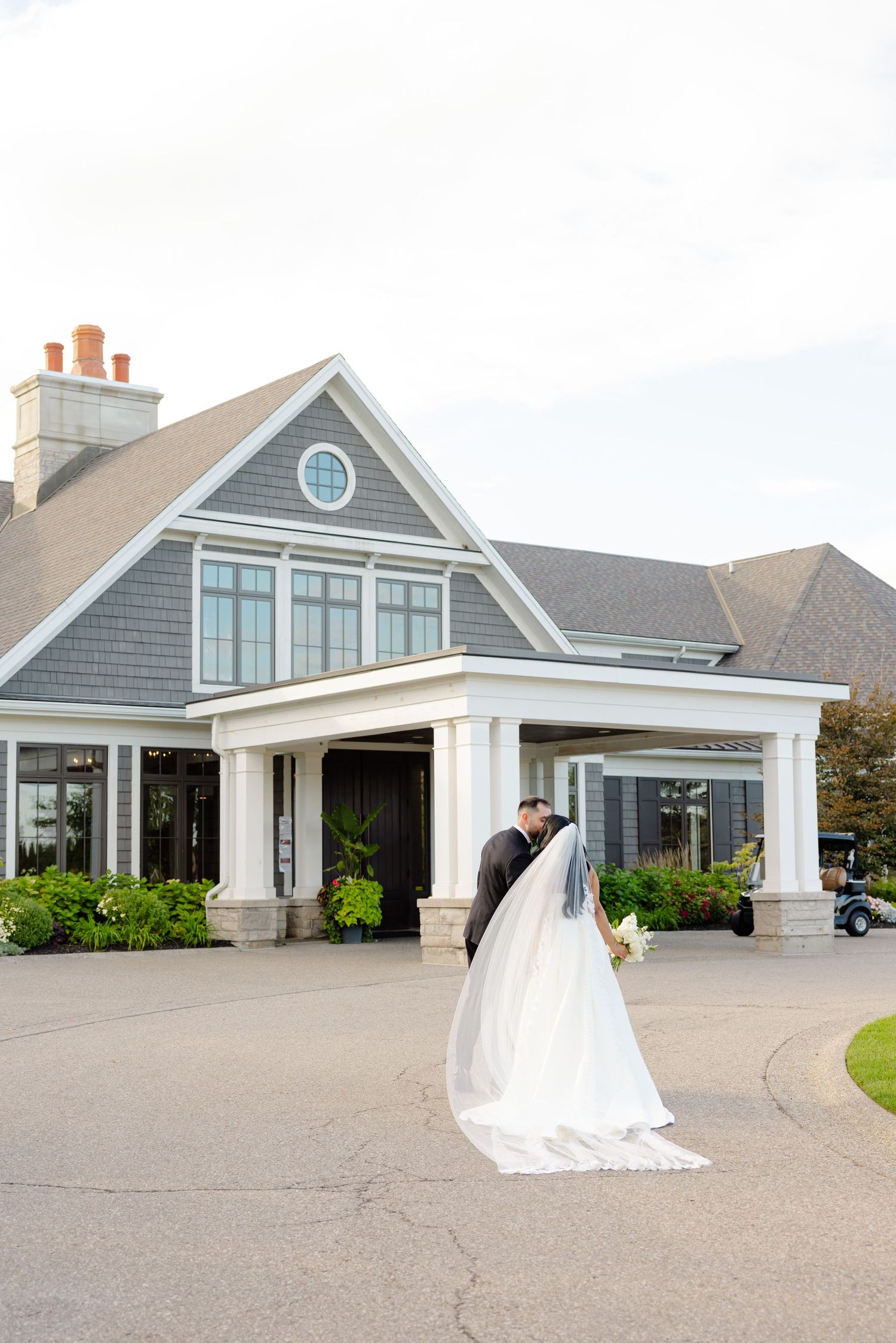 A wedding couple enters a gray coastal-style house with white columns and a circular window.