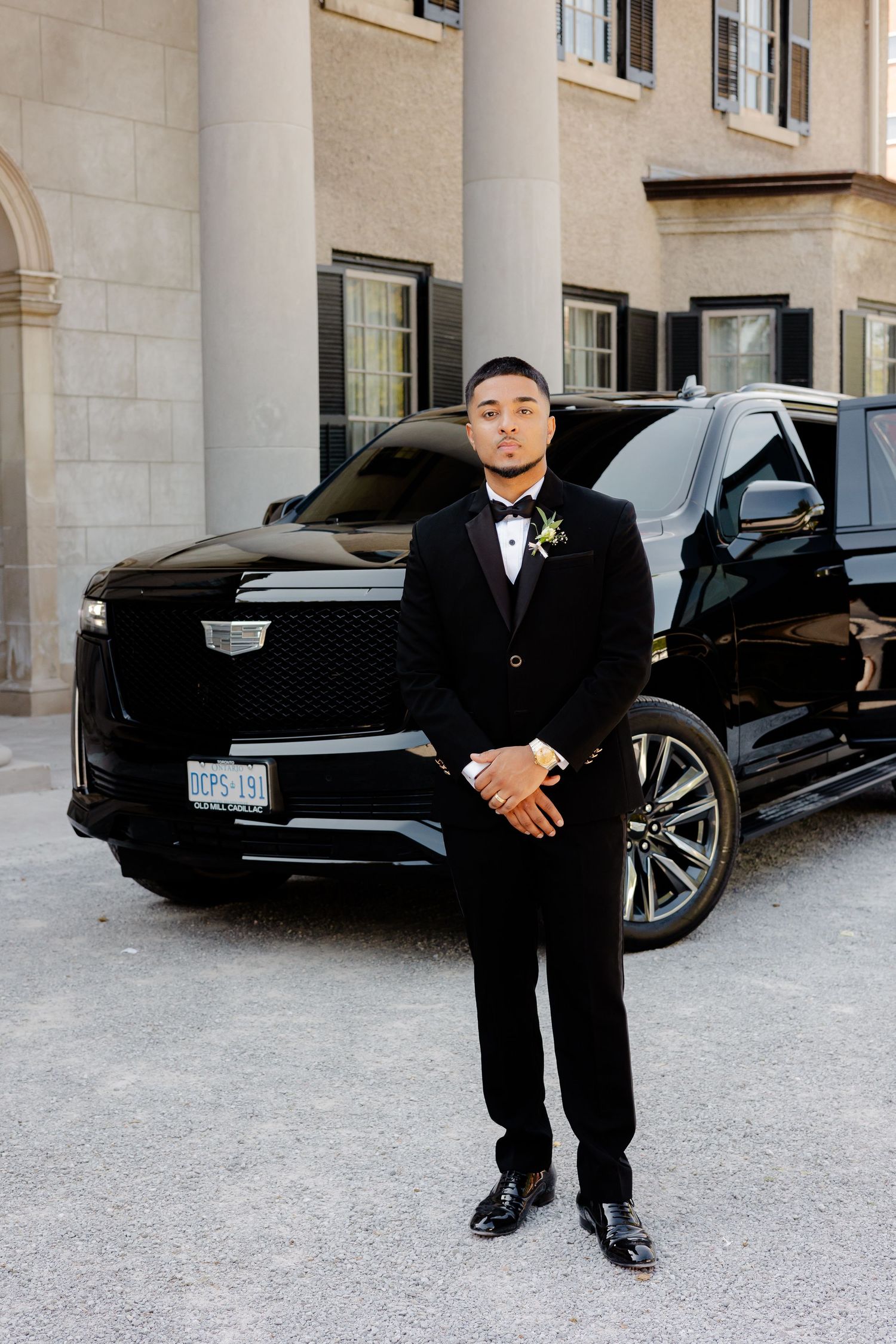 Groom in classic tuxedo poses in front of a black Cadillac SUV at an elegant venue.