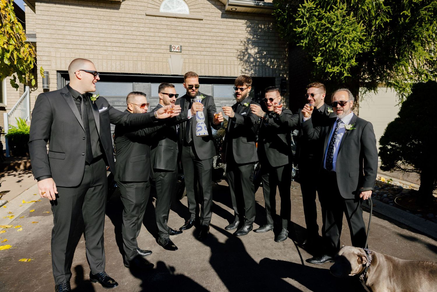 Groomsmen in dark suits and sunglasses pose together for a fun group photo outdoors.