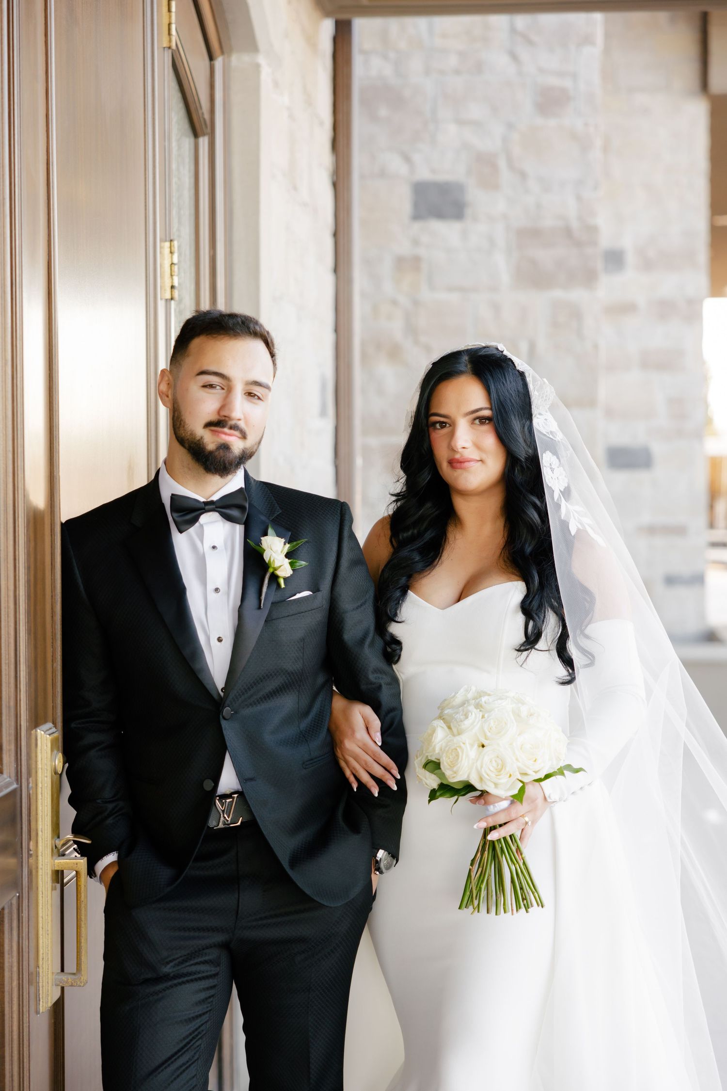 Classic wedding portrait of couple with white floral bouquet against stone wall.