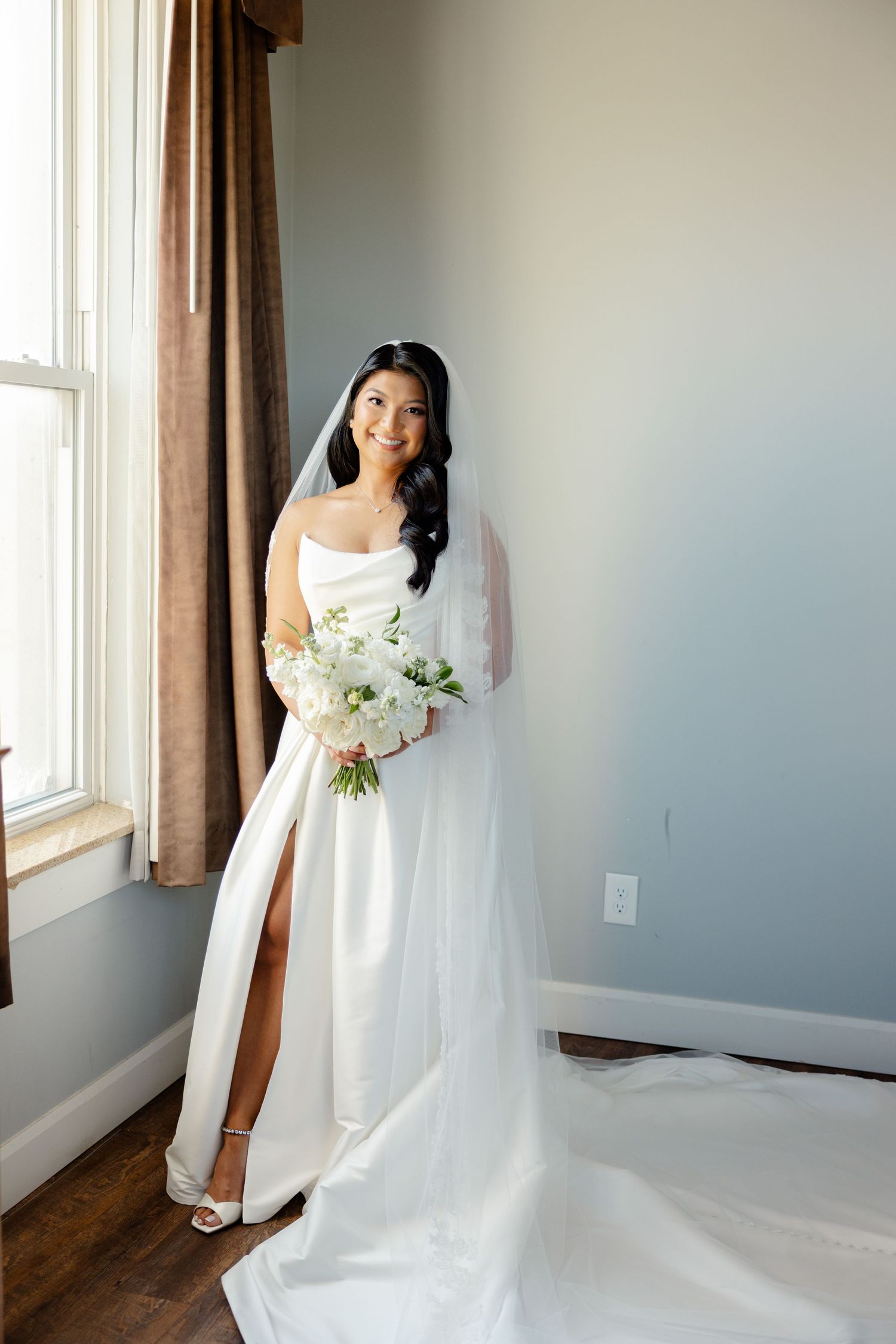 Full-length portrait of bride in modern wedding dress with leg slit and flowing veil by window.