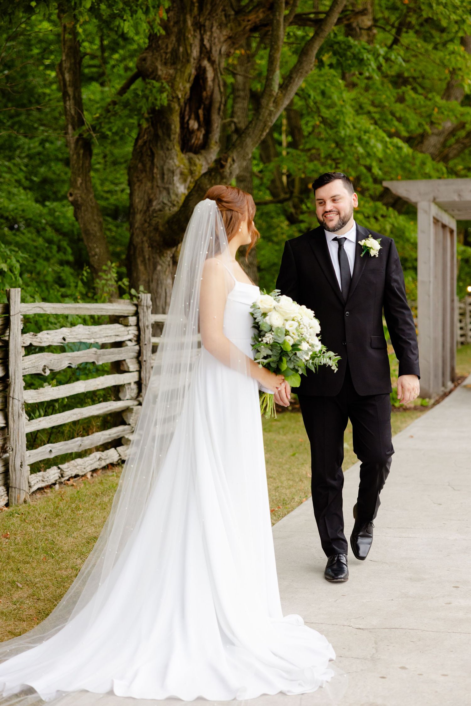 Couple in wedding attire walking together on outdoor path surrounded by trees and rustic fencing.