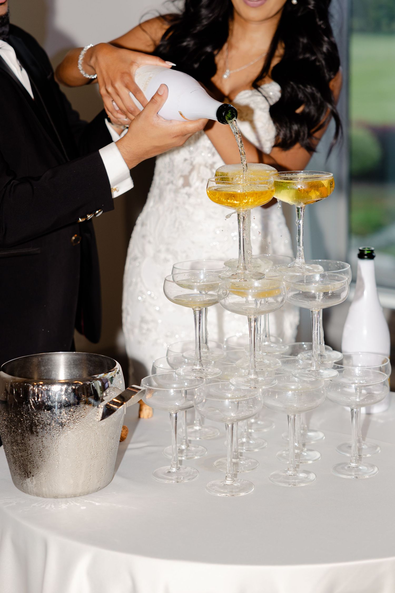 A champagne tower being set up at a wedding reception with glasses and an ice bucket on a white tablecloth.