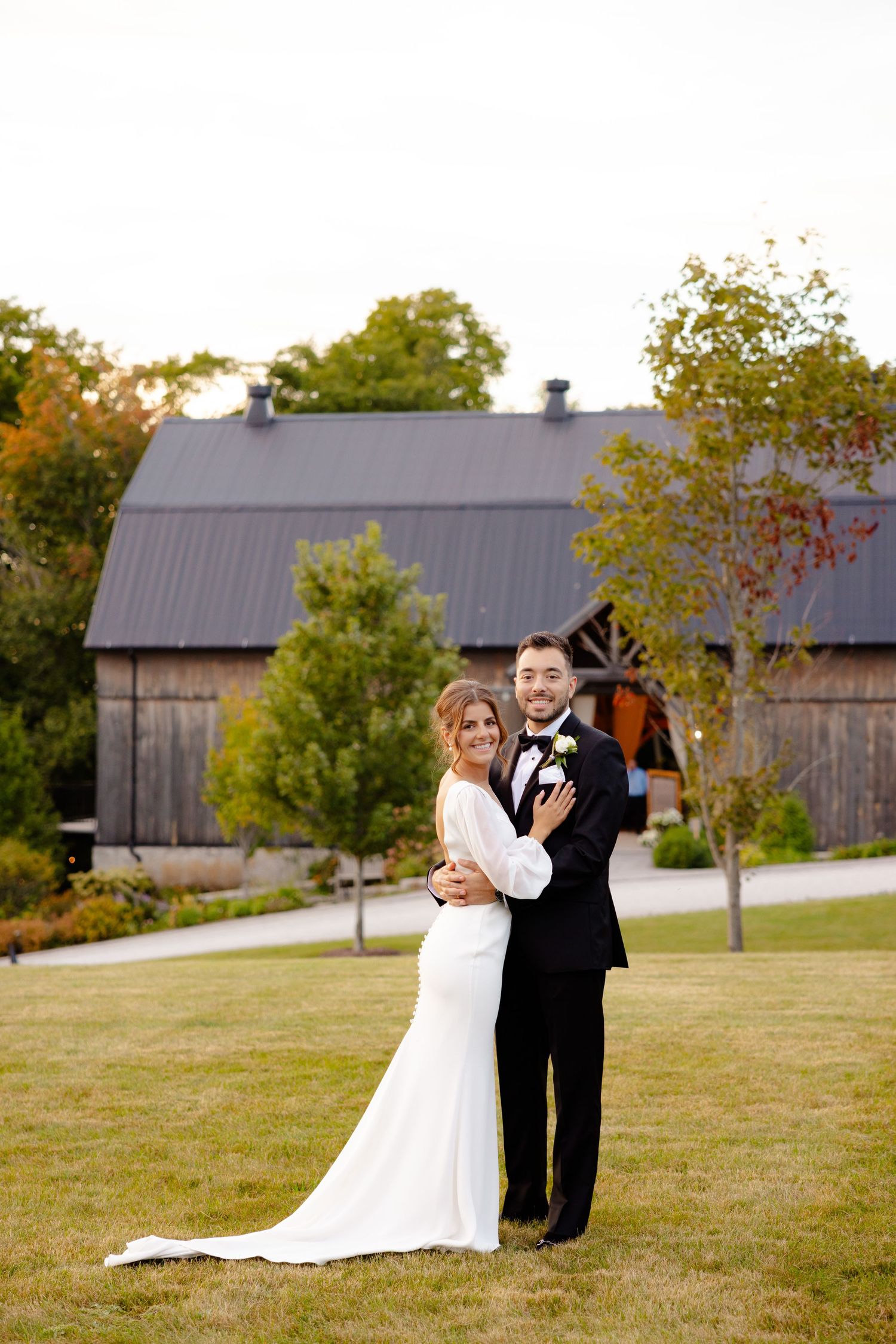 A wedding couple poses in formal attire in front of a rustic barn during golden hour.