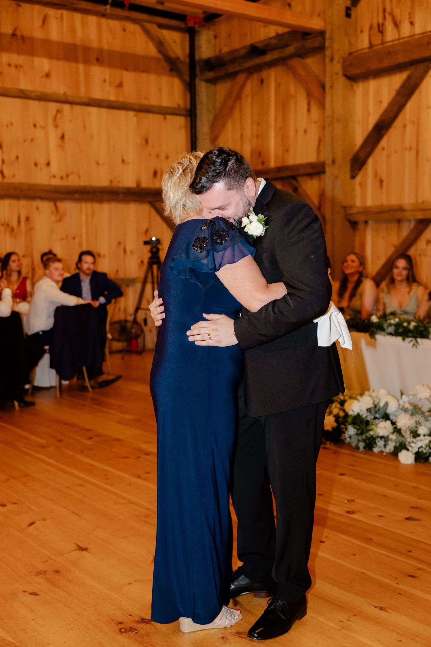 Two people embrace during a dance at a barn wedding with wooden walls and elegant table settings.