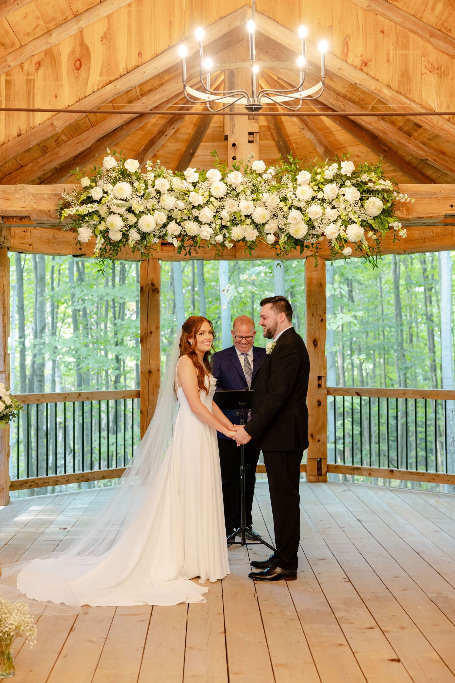 Wedding couple exchanging vows under elegant white floral arrangement in timber-frame pavilion.