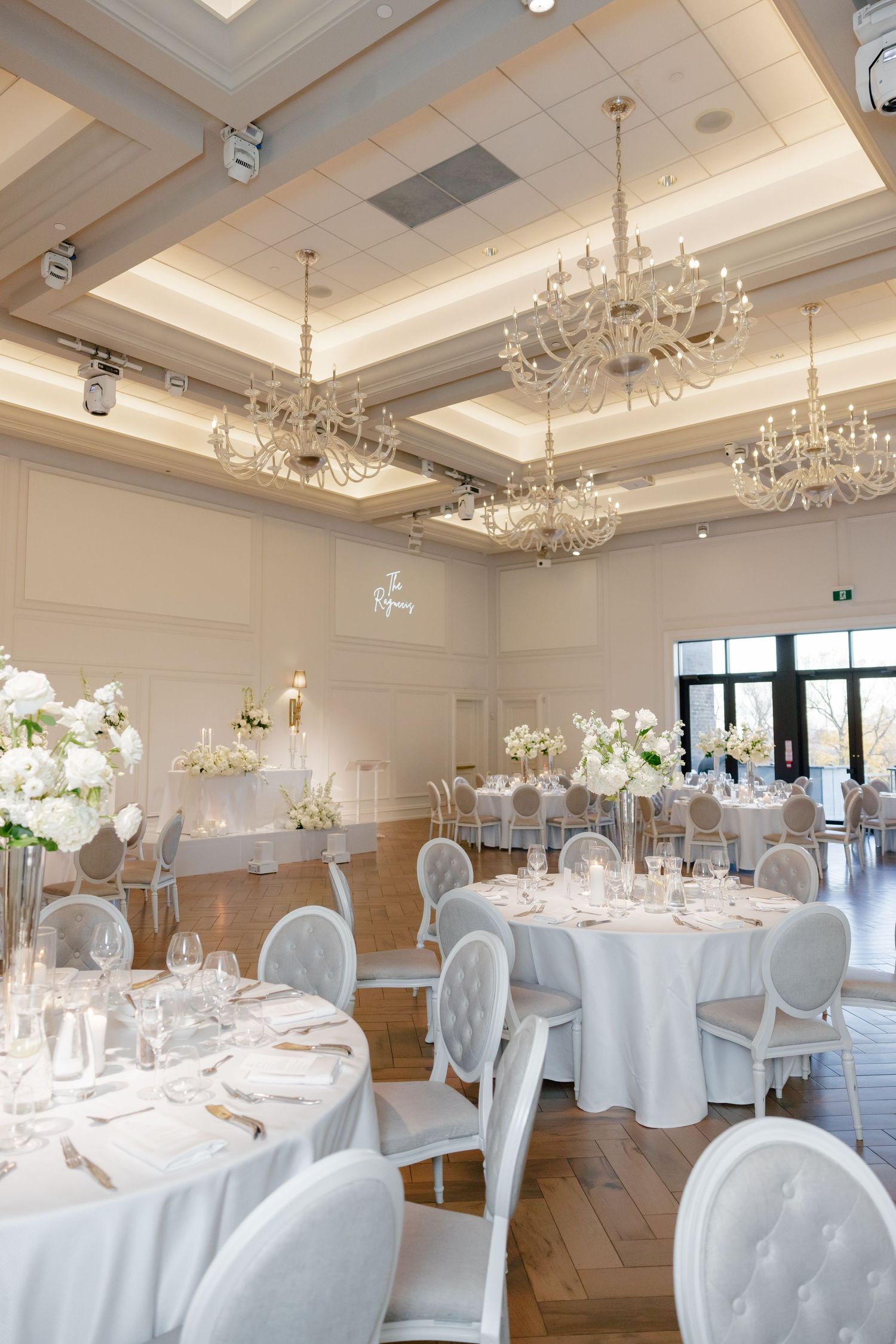 Elegant ballroom with herringbone floors, crystal chandeliers, and white round tables set for a wedding.