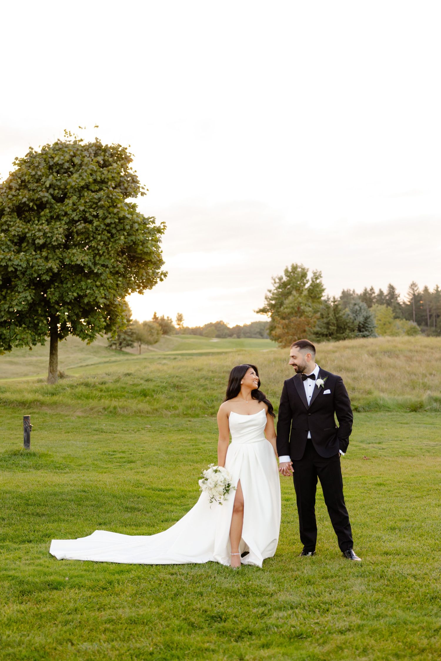 Newlyweds stand together on a scenic hillside with a lush green tree and forest backdrop.