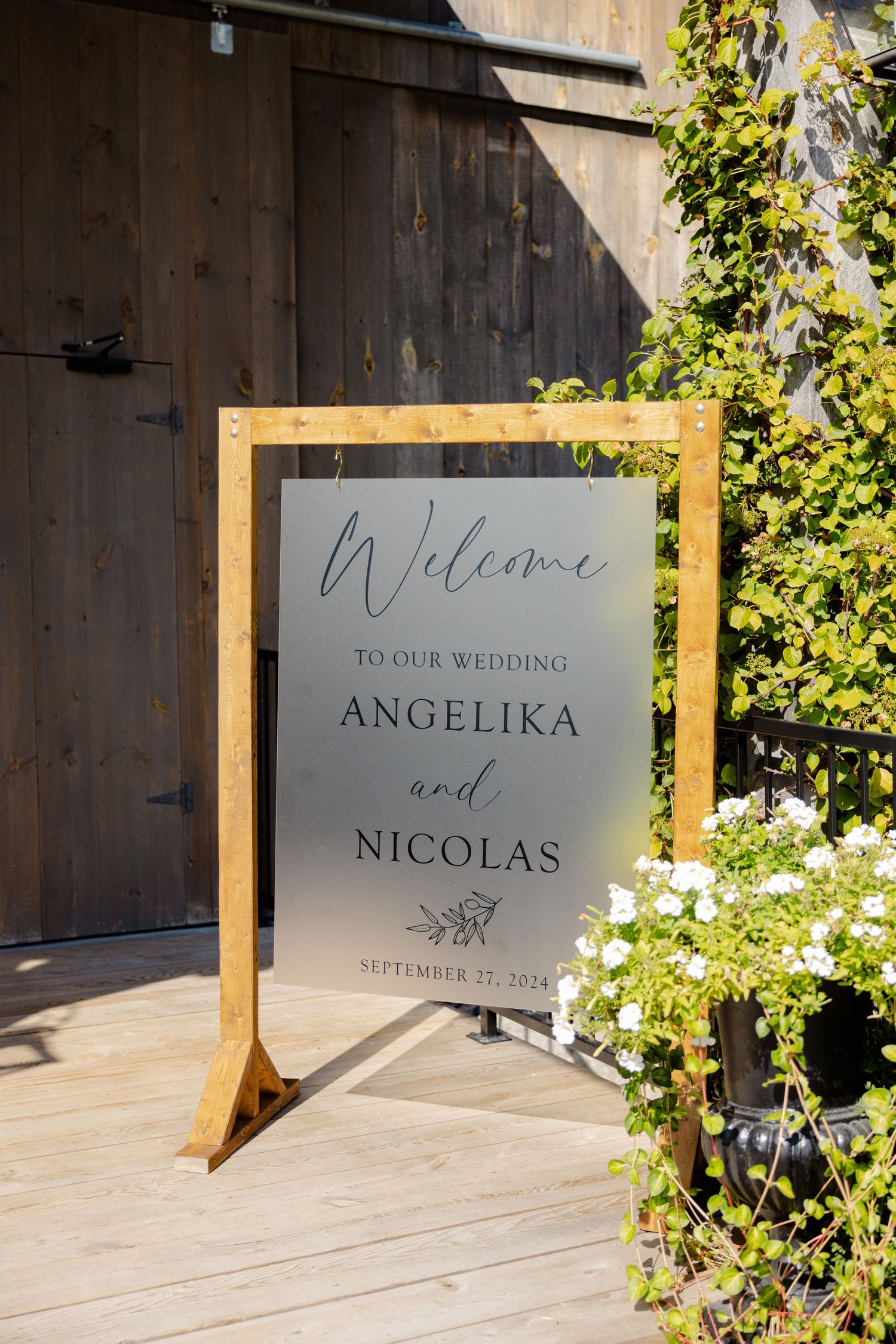 A wooden-framed wedding welcome sign for Angelika and Nicolas displayed against a rustic barn wall.