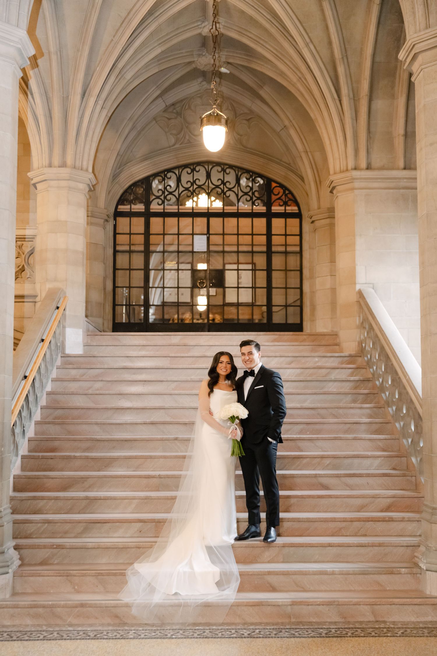 Couple in wedding attire poses on grand marble staircase beneath gothic archway and vintage lighting.