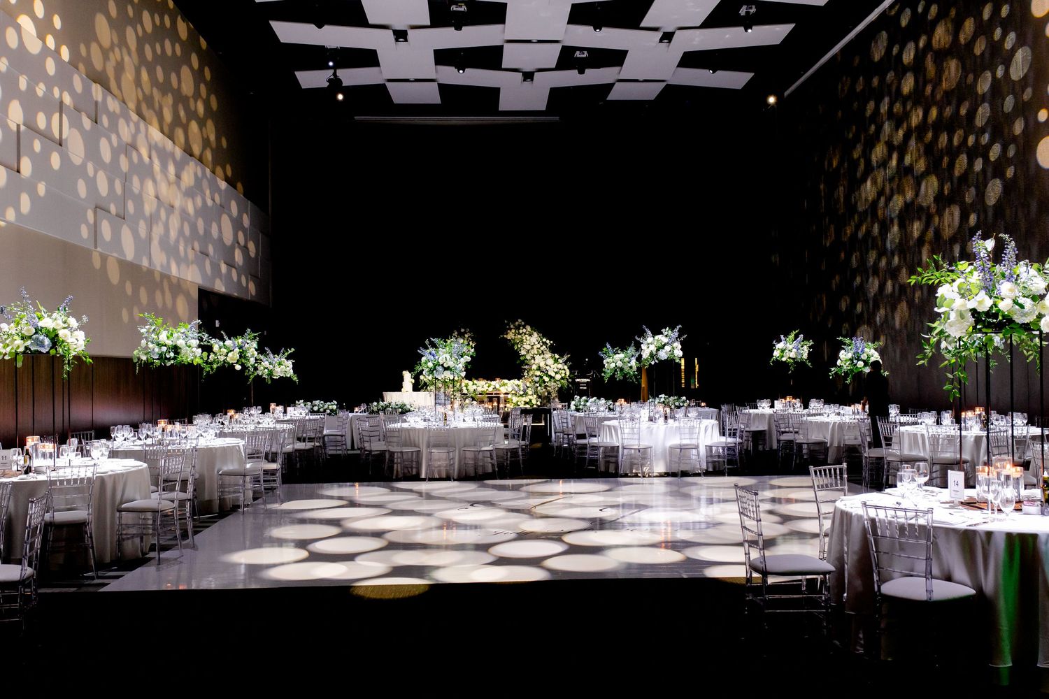 Modern wedding venue with dramatic spotlight effects and floral centerpieces on white-clothed tables.