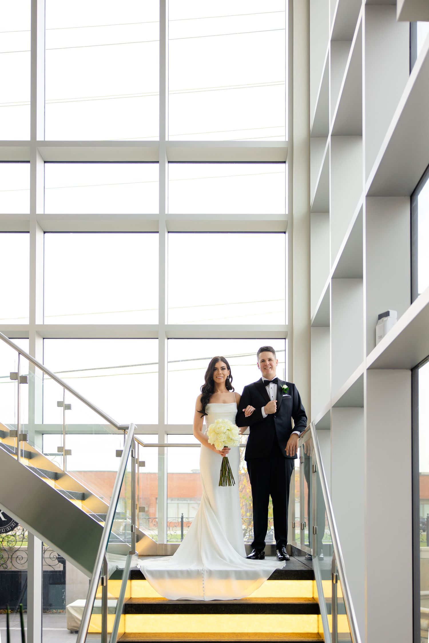 Elegant couple poses on architectural staircase with floor-to-ceiling windows.