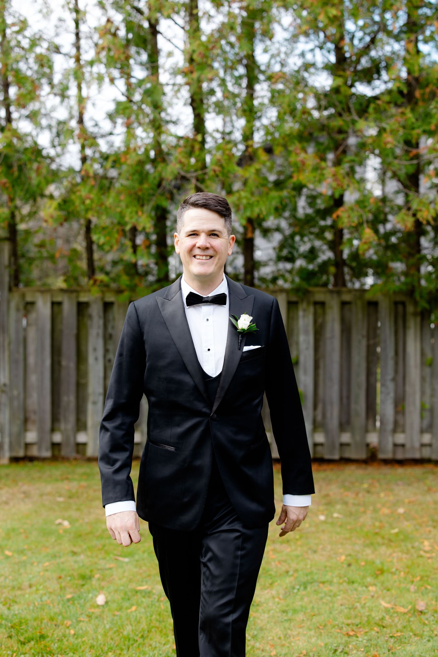 Wedding portrait of a groom in black tuxedo with boutonniere walking outdoors in front of a wooden fence.