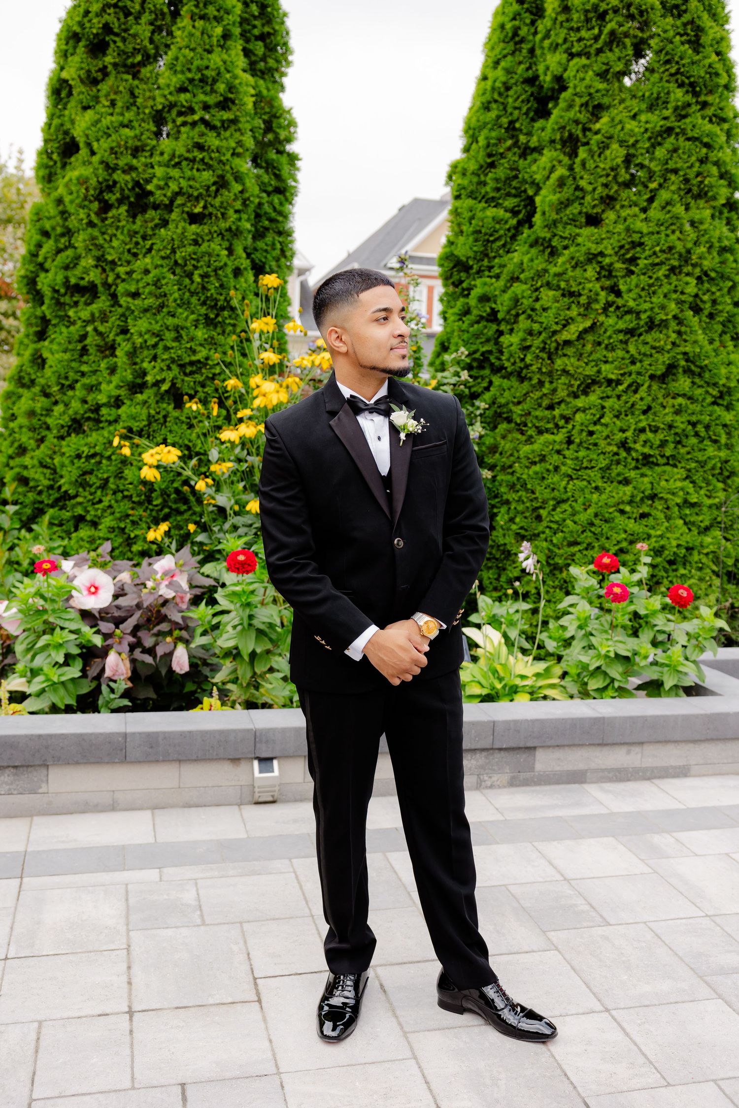A groom in a classic black tuxedo poses elegantly in a garden with tall evergreen trees and colorful flowers.