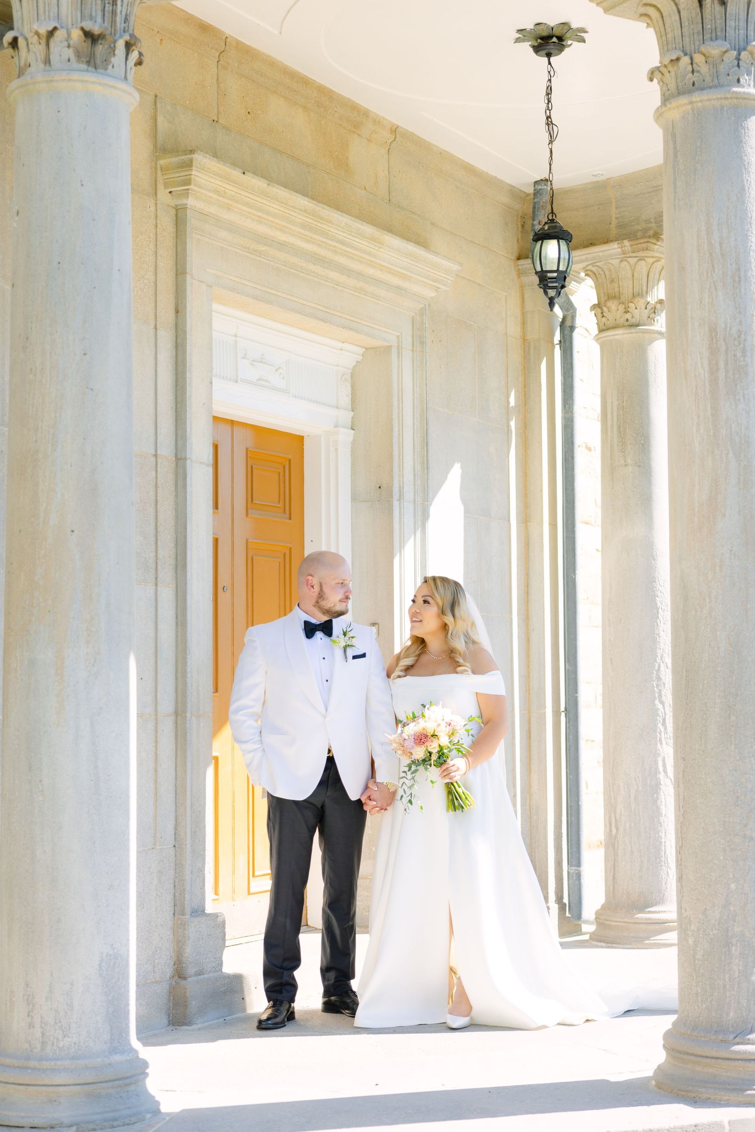 Wedding couple poses between ornate columns with hanging lantern in classic architectural setting.