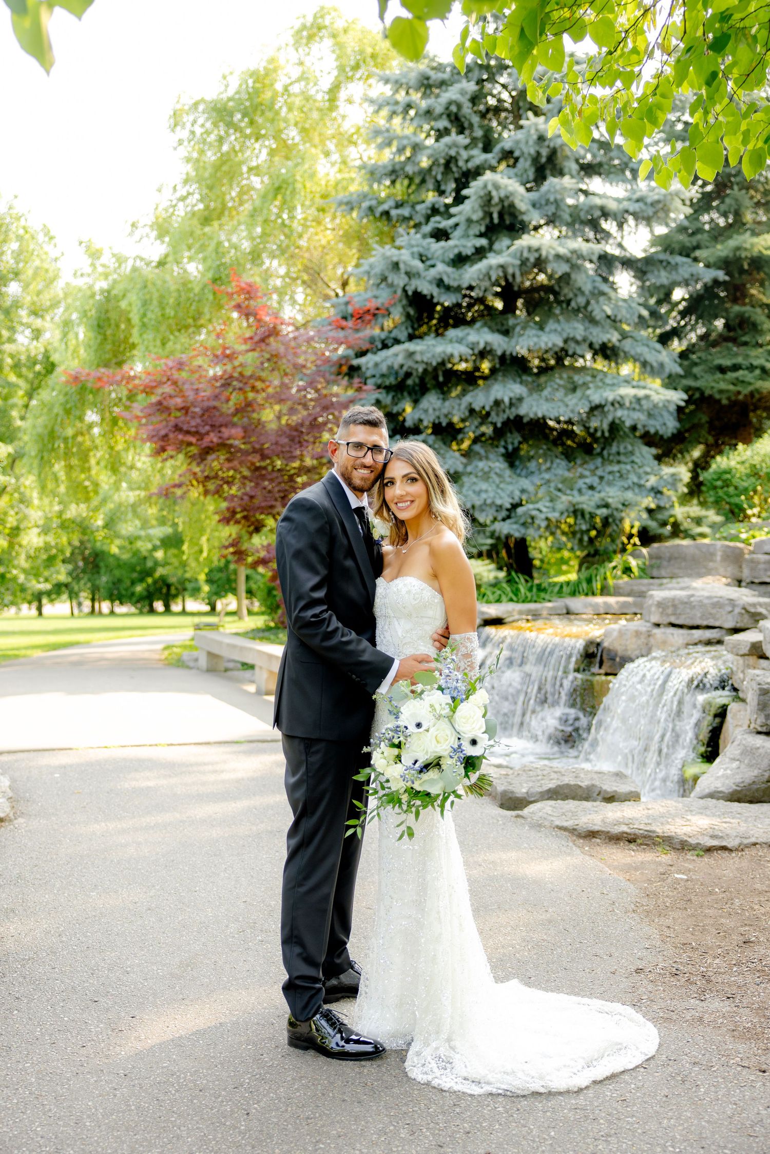 A couple in formal wedding attire poses in front of a waterfall and evergreen trees on their wedding day.