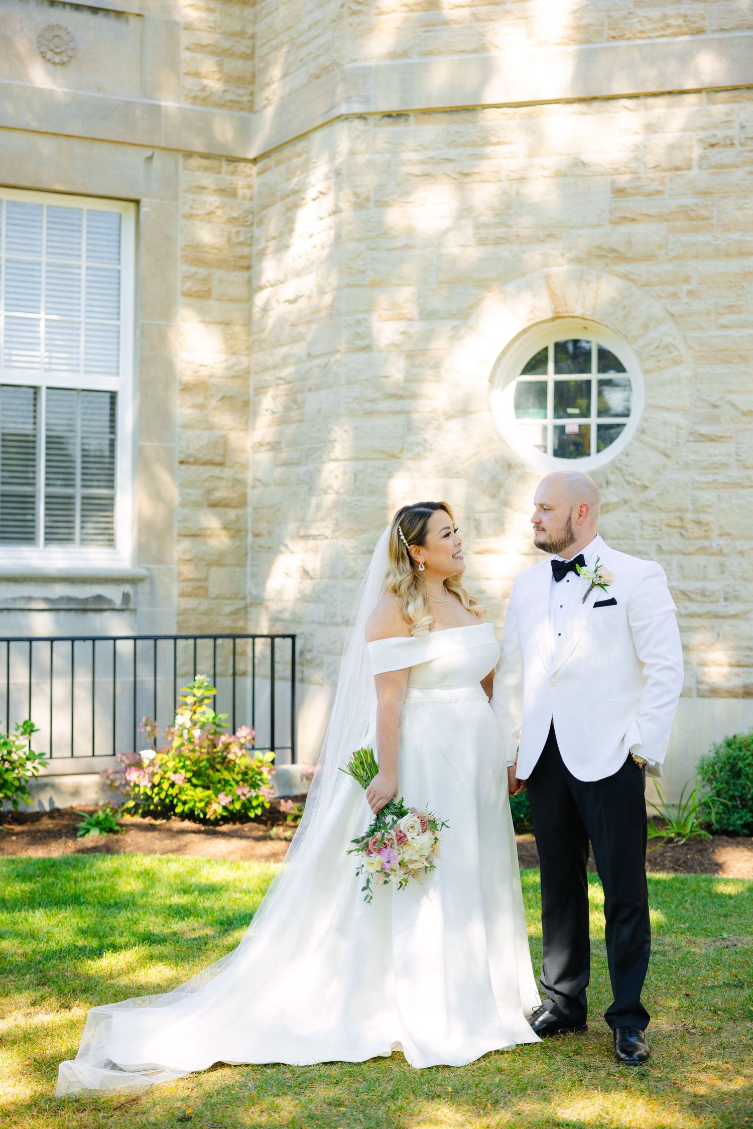 A wedding portrait in front of a limestone building showing an elegant couple in white formal attire.
