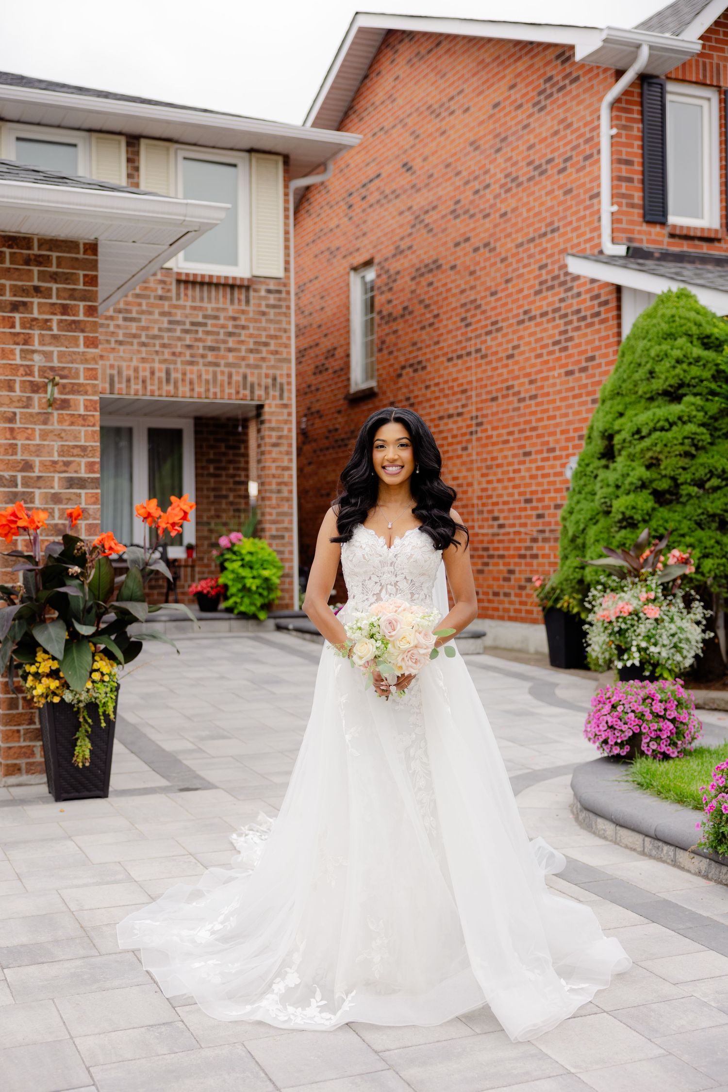 Radiant bride in flowing white dress poses in front of brick home with colorful garden.