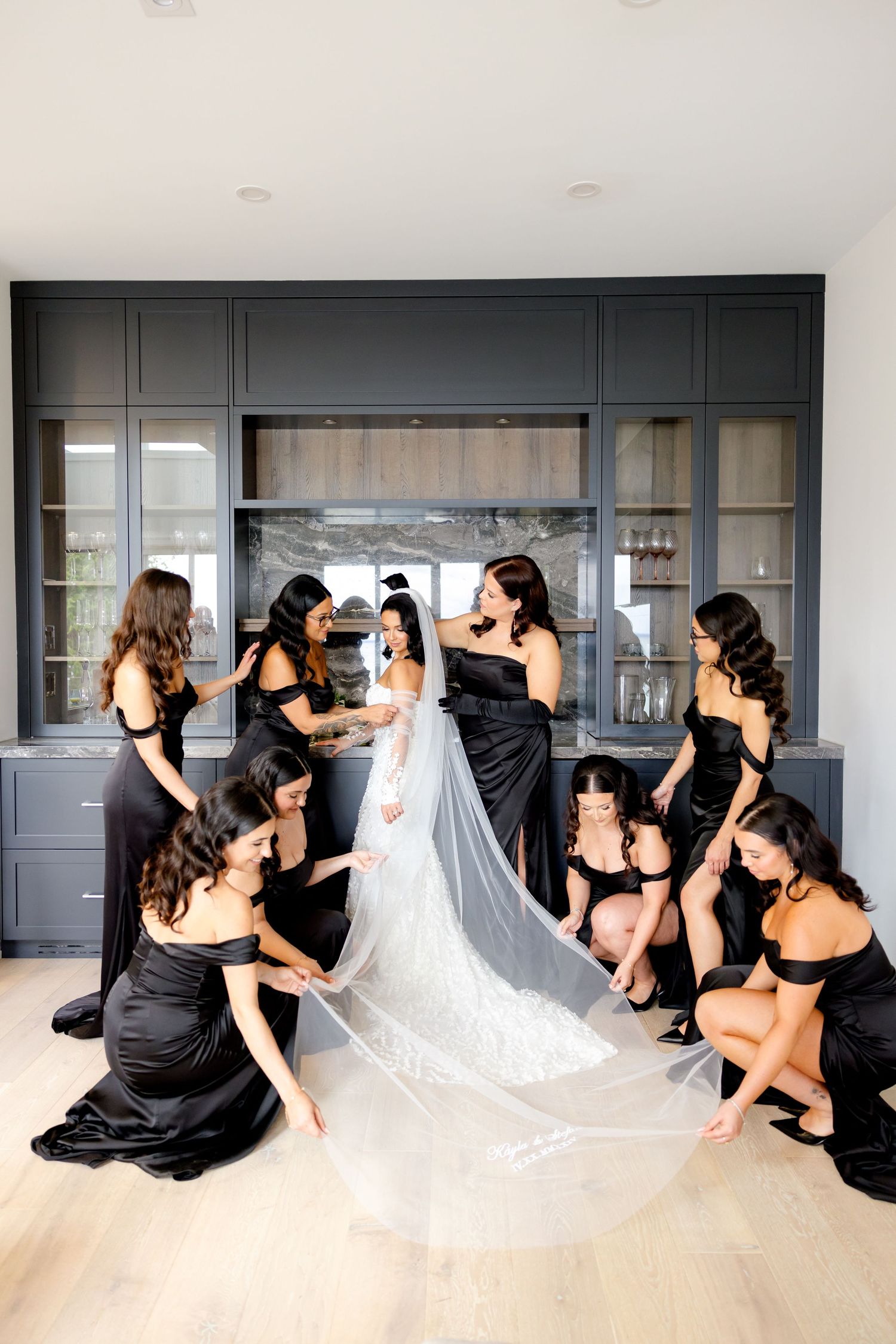 Bridesmaids gather around to help arrange bride's long veil in modern interior setting.