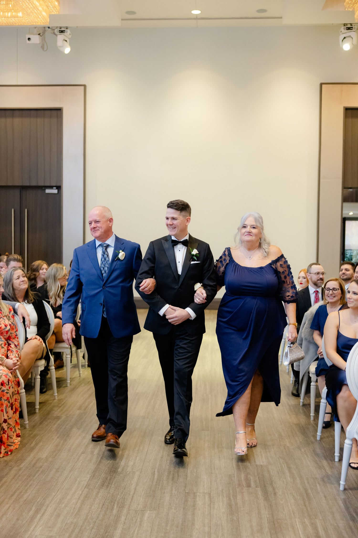Wedding processional with family members walking down the aisle in formal navy and black attire.