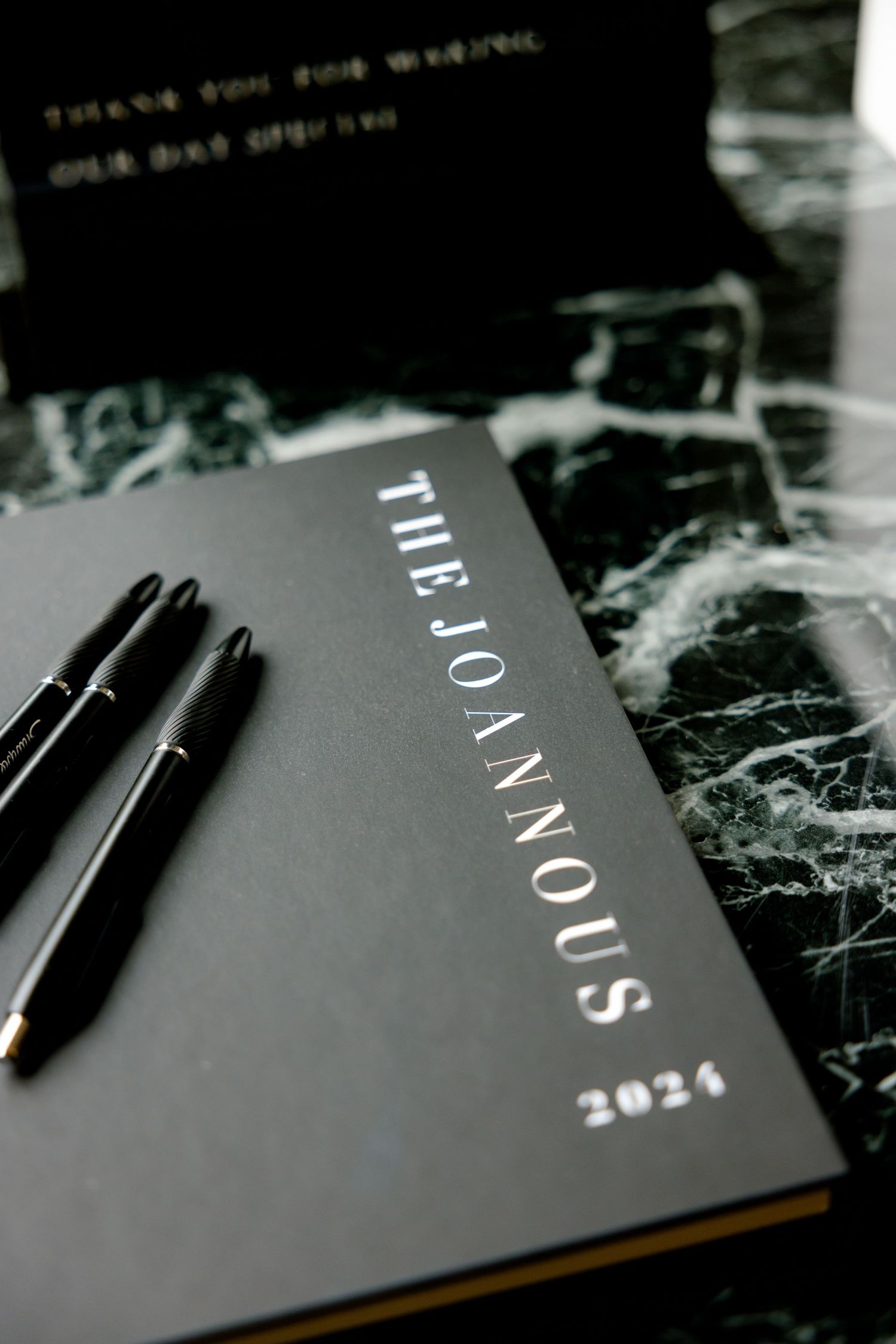 Wedding planning book titled The Jonnous 2021 with black pens on marble surface.