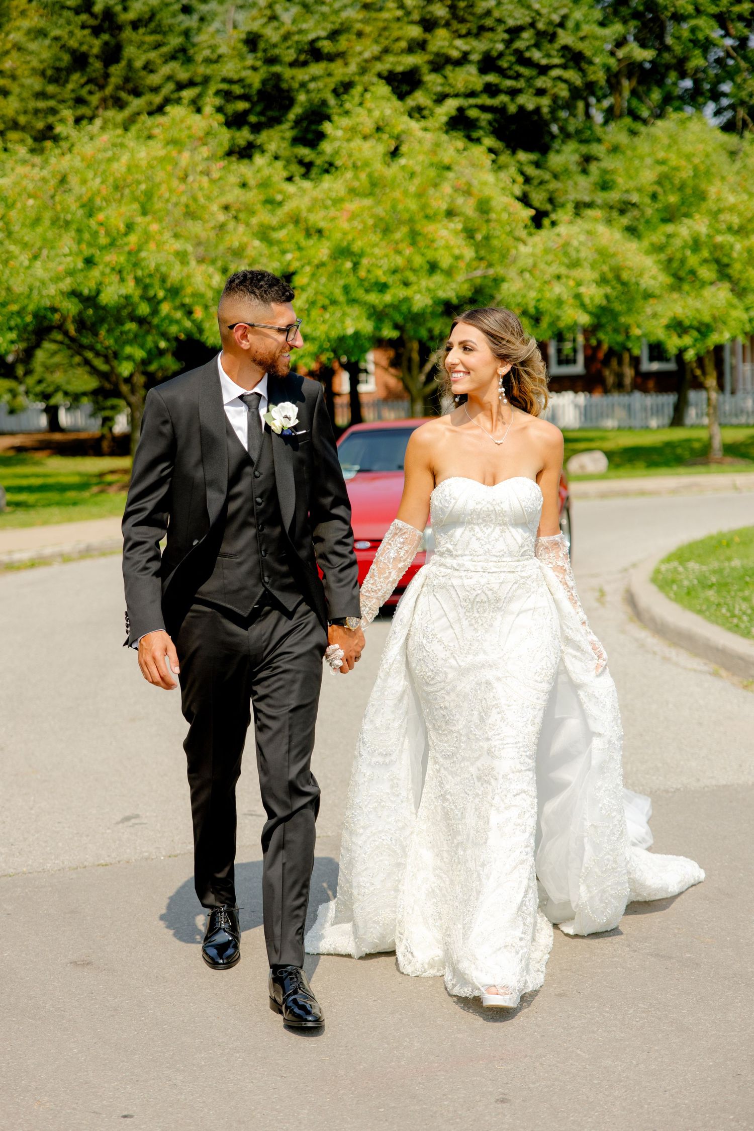 Joyful newlyweds walk together on a curved path with trees and a red car behind them.