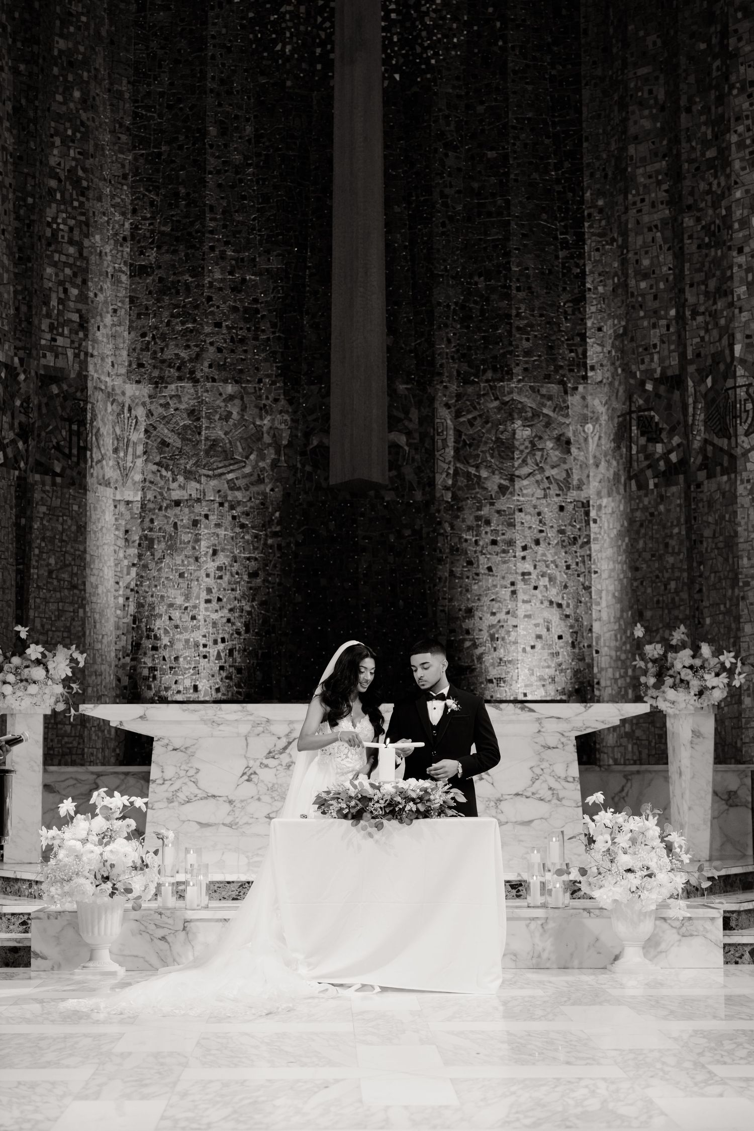 Black and white photo of newlyweds signing documents at marble altar with floral arrangements.