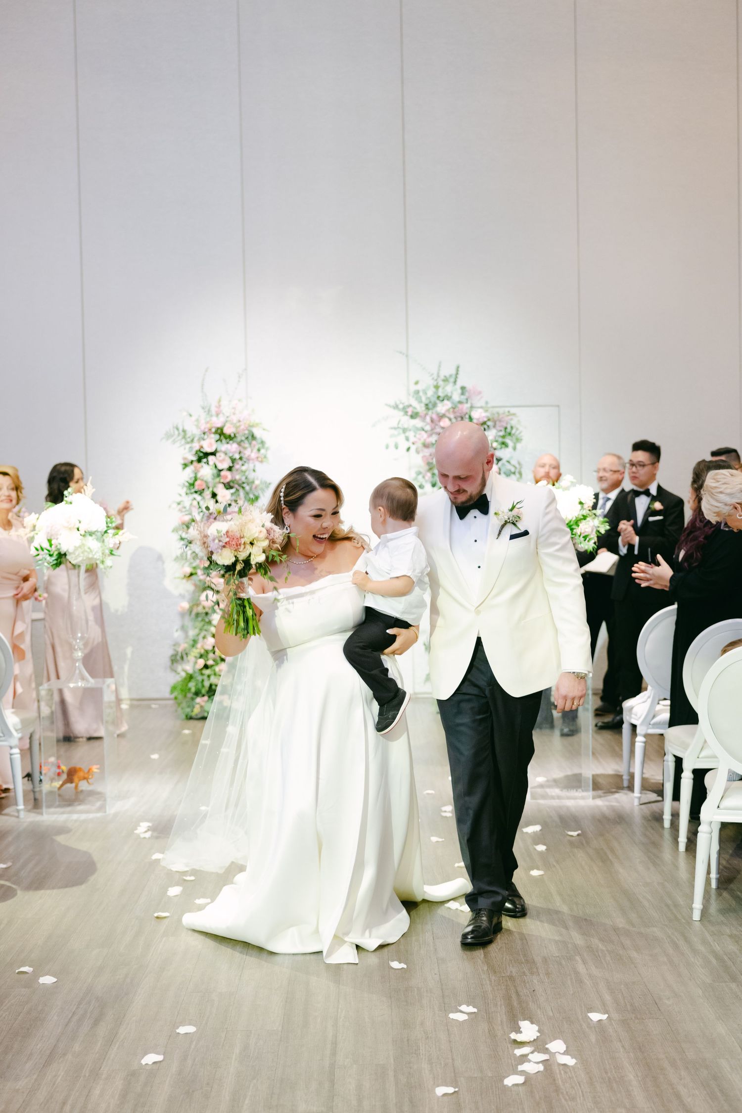 Joyous newlyweds walk down the aisle with a small child during their elegant indoor wedding ceremony.