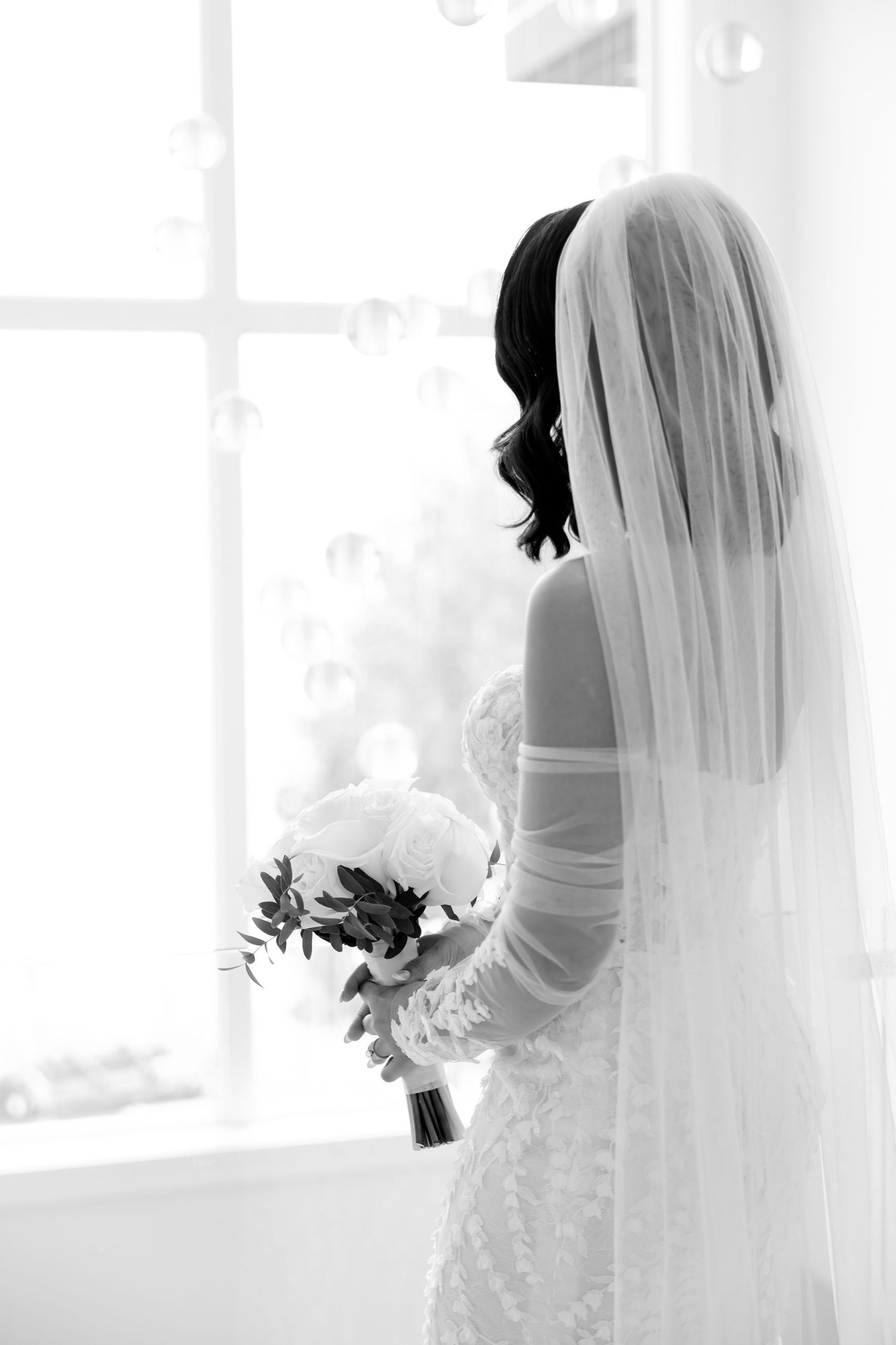Black and white artistic bridal portrait with flowing veil and bouquet near window.
