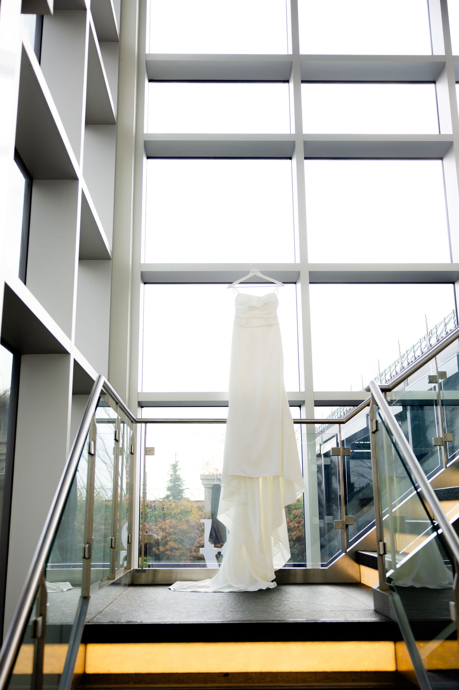 Elegant white wedding dress hanging in a modern stairwell with floor-to-ceiling windows.