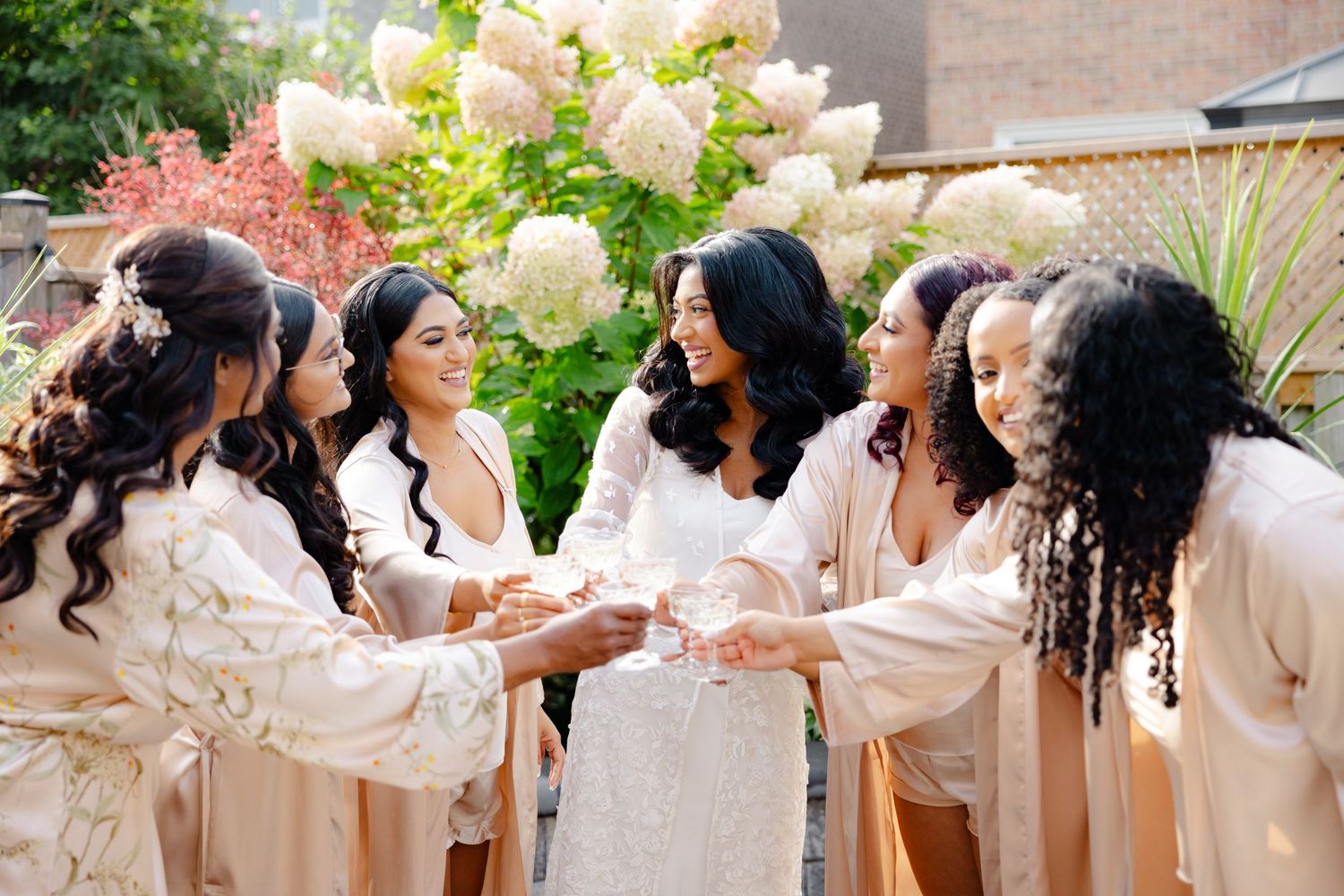 Wedding party toasts together in matching champagne-colored robes in a garden setting with blooming hydrangeas.