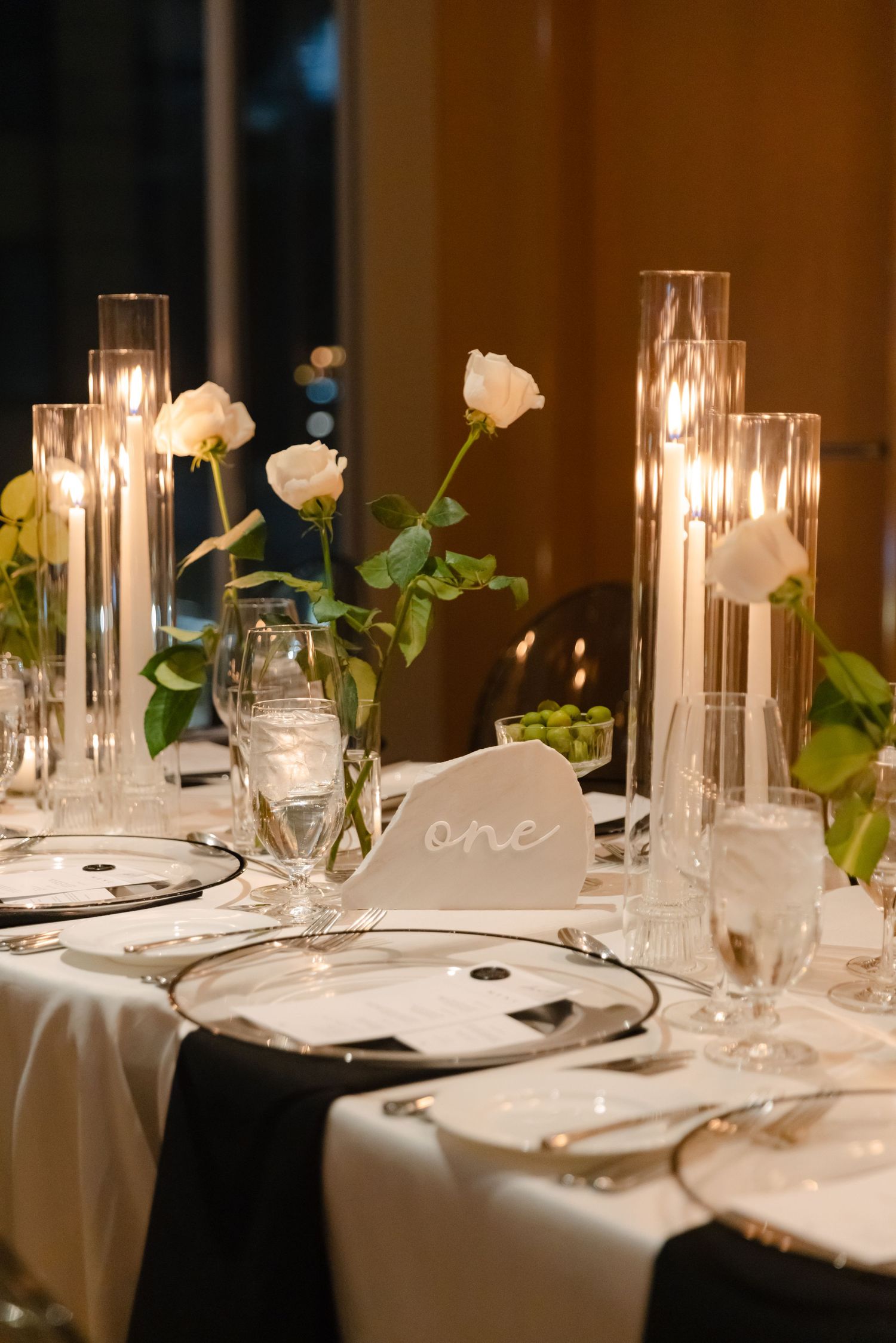 Wedding table decor featuring white roses, glowing candles in glass cylinders, and elegant place settings.