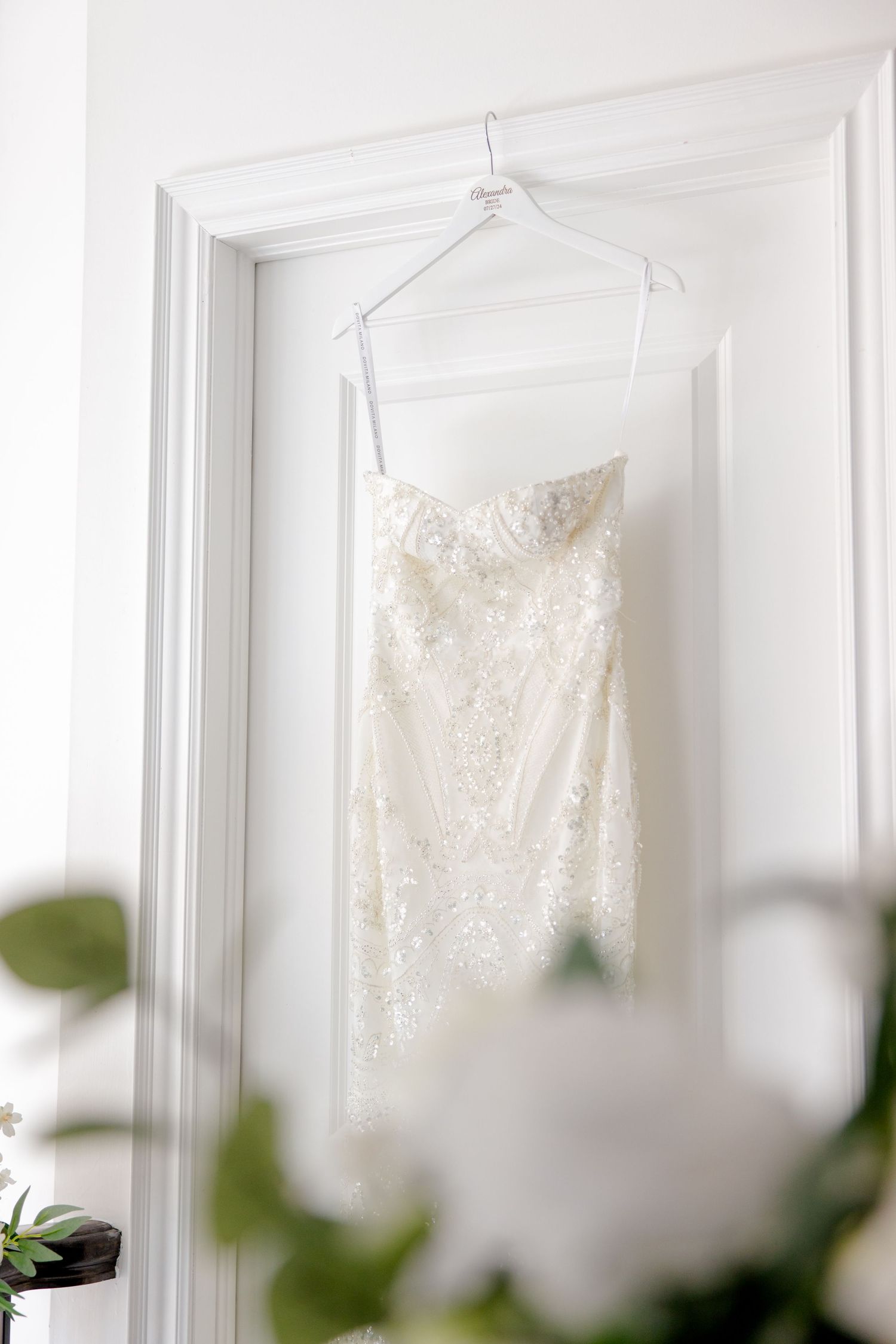 An elegant white lace wedding gown displayed on a hanger against a bright white doorframe.
