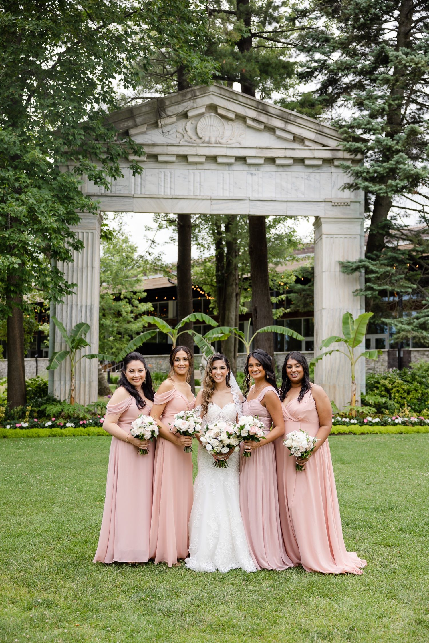 Wedding party in blush pink dresses poses under classical white stone archway in garden setting.