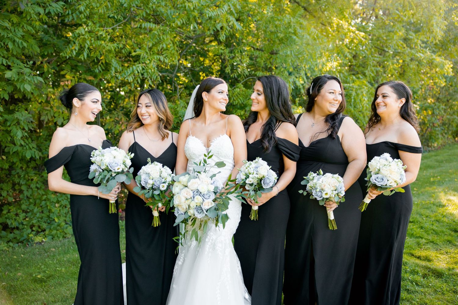 Wedding party poses in black dresses and white gown against green foliage background.