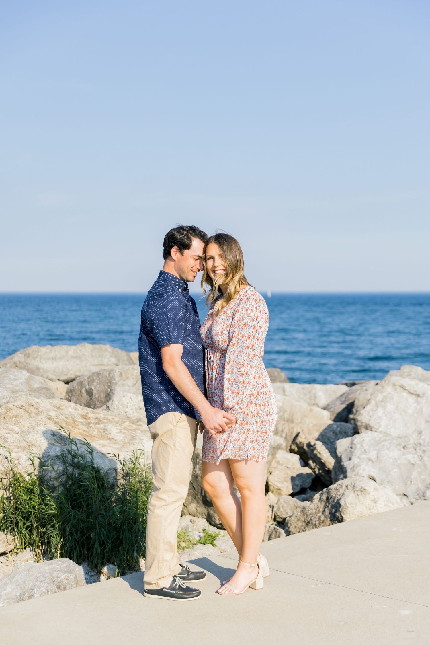 A romantic couple standing close together on a rocky beach with blue ocean backdrop.