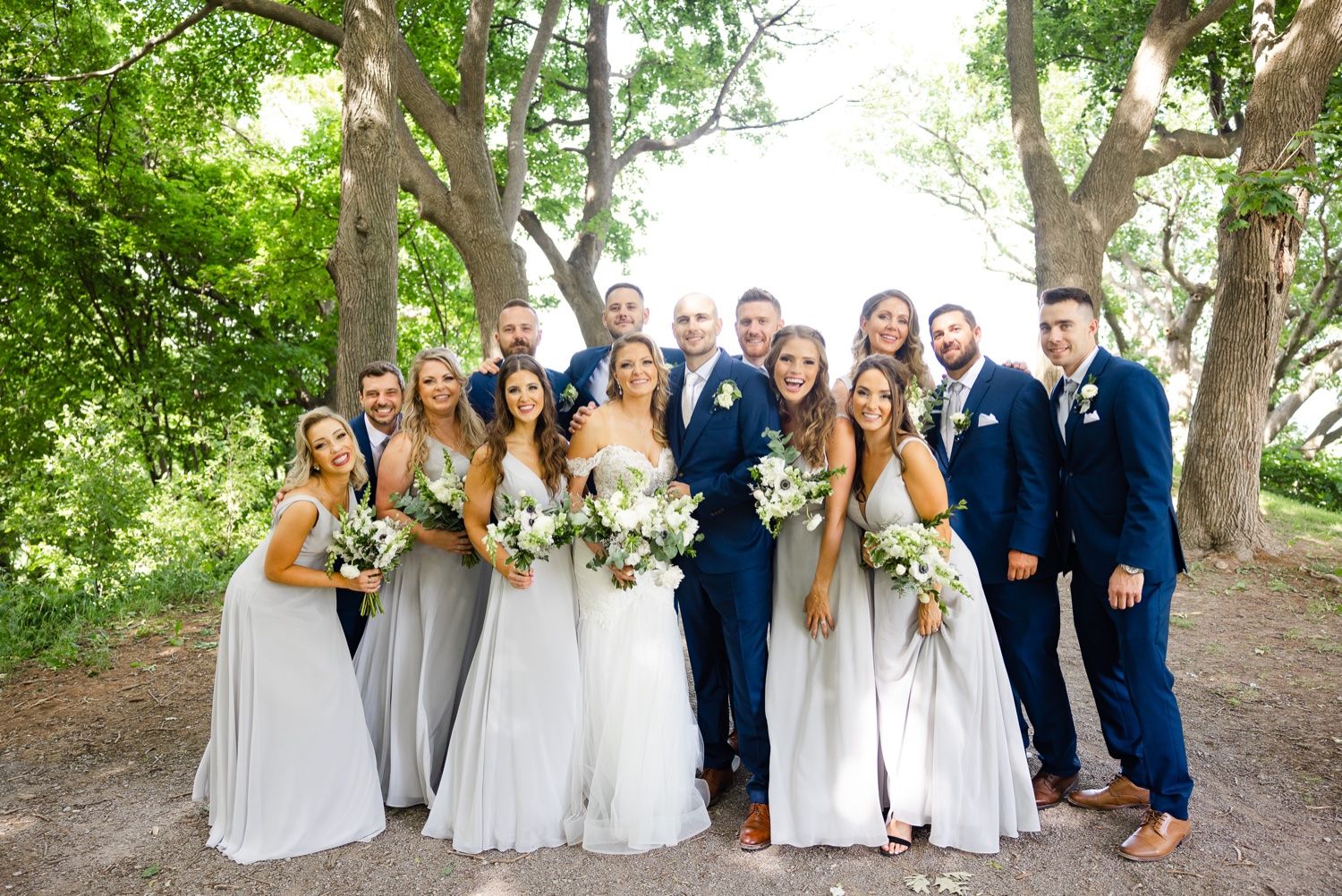 Full wedding party in navy suits and light gray dresses poses together under a canopy of trees.