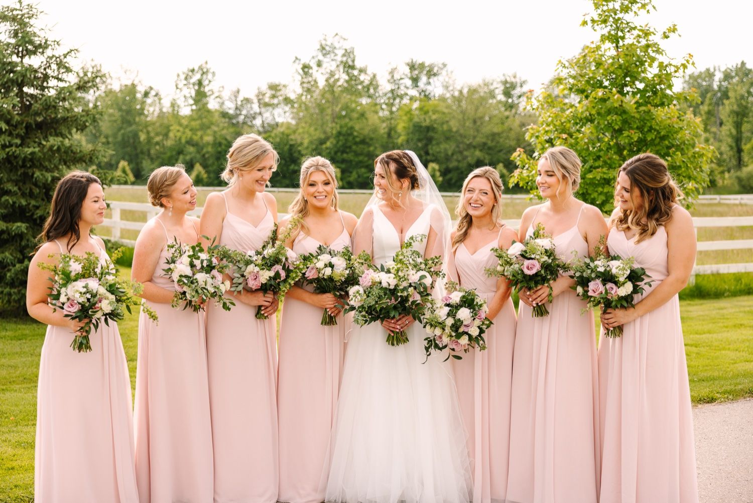 Bridesmaids in blush pink dresses stand together holding floral bouquets at an outdoor wedding venue.