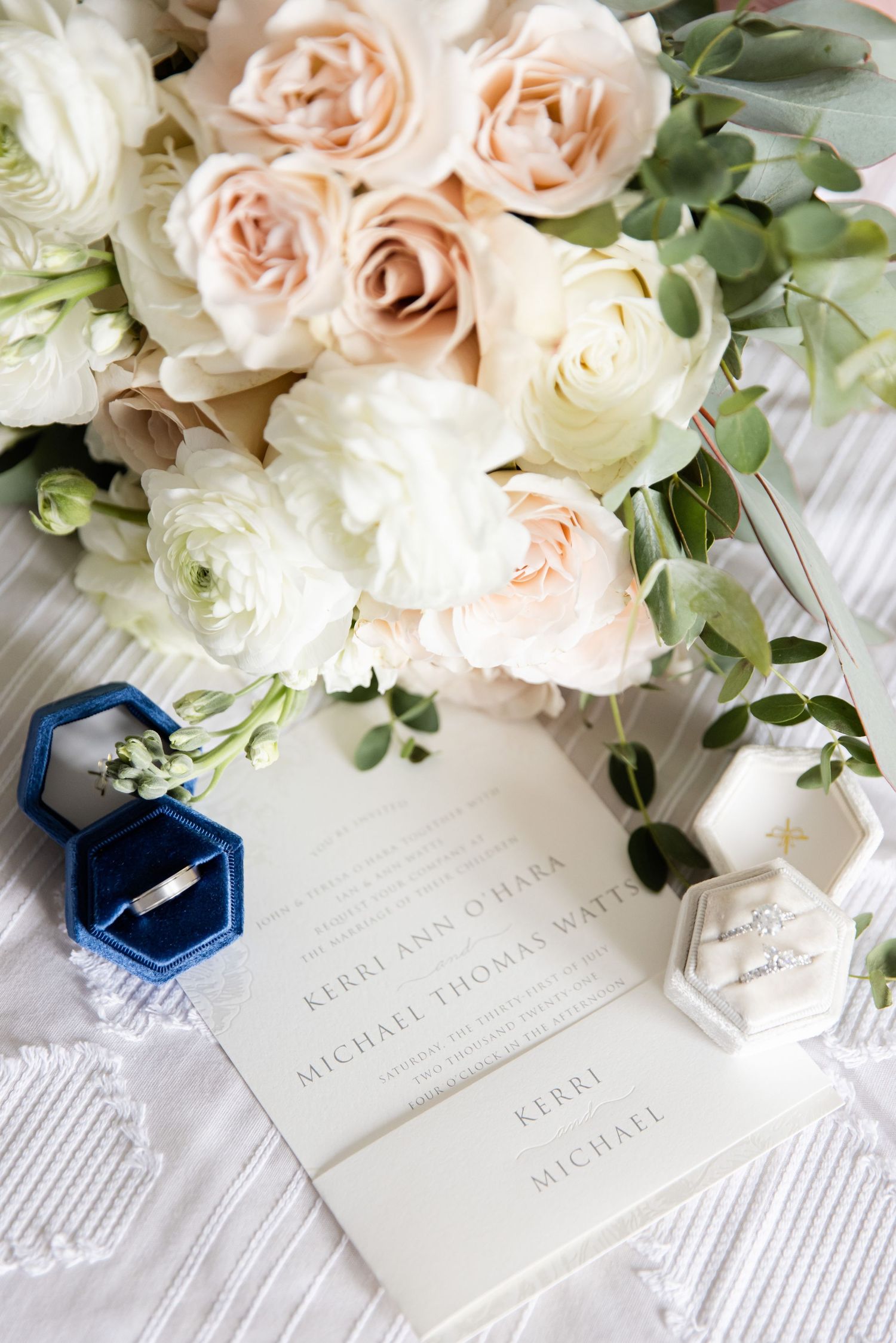 Romantic blush and white rose bouquet with blue ring box and wedding stationery on white textured surface.