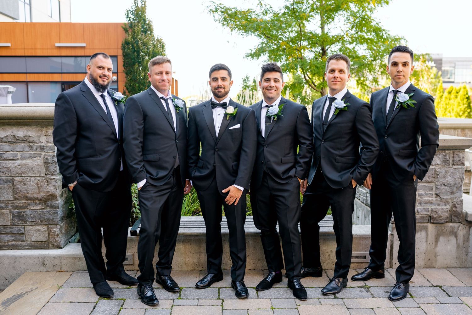 Groomsmen in black suits and ties pose together on stone wall with urban backdrop.