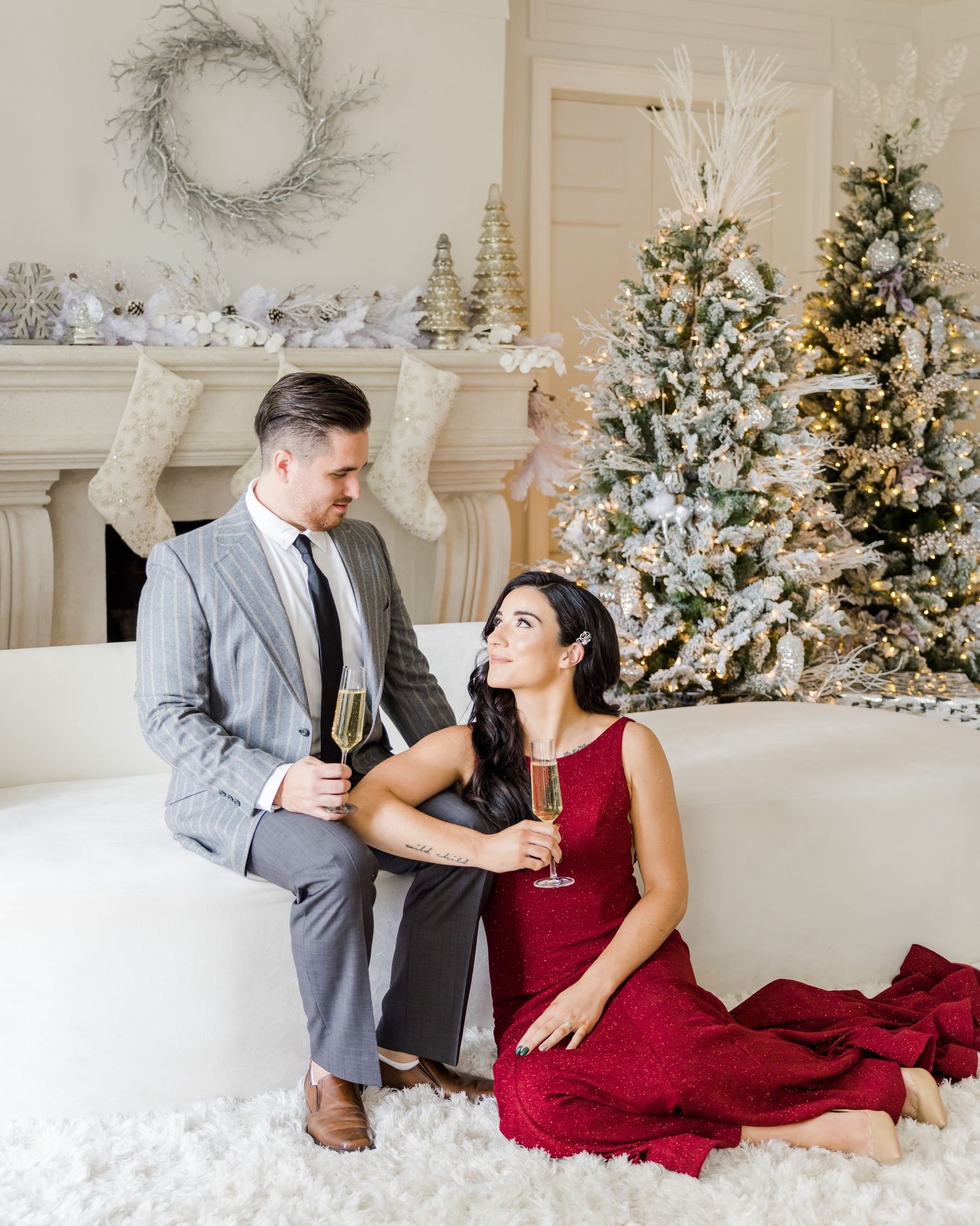 Couple sharing champagne while seated in a festive white room with decorated Christmas trees.