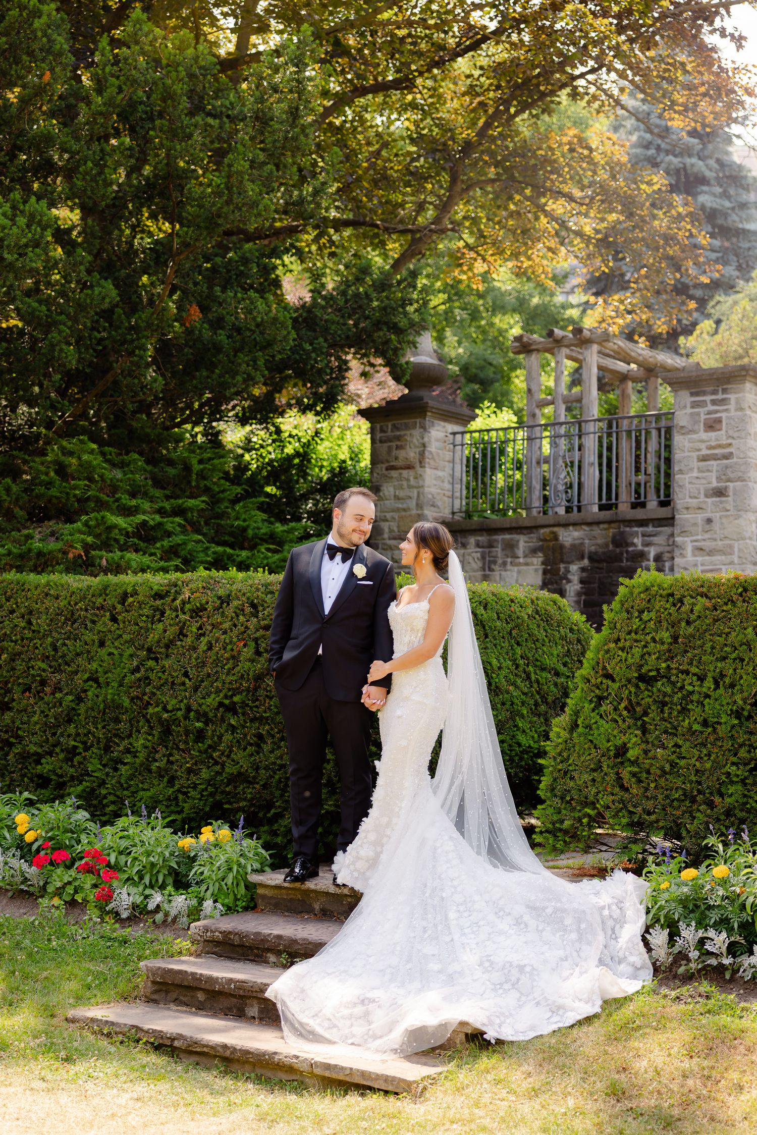 Wedding couple stands on stone steps in a lush garden setting with blooming flowers and manicured hedges.
