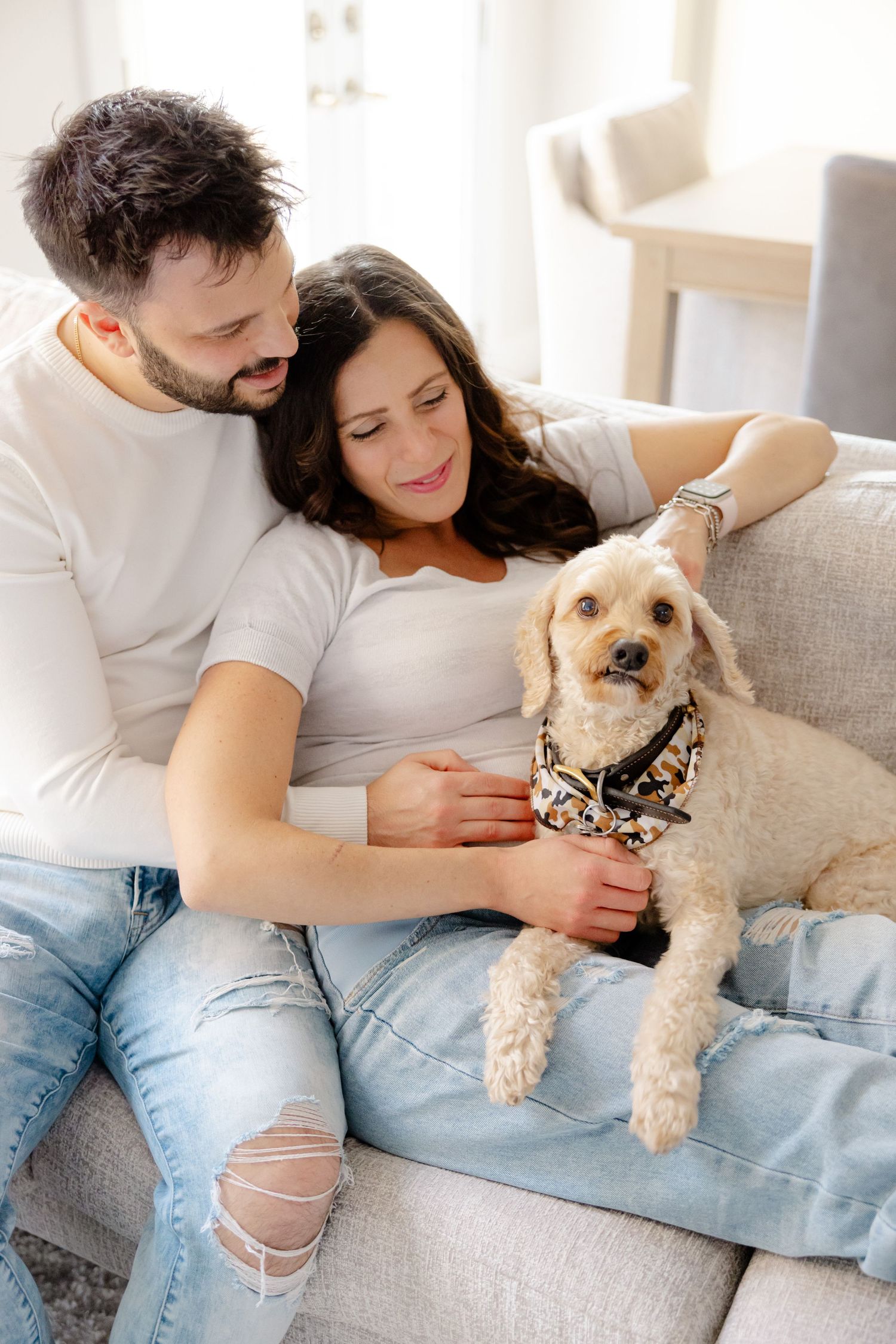 A couple snuggles on a couch with their small dog wearing a bandana.