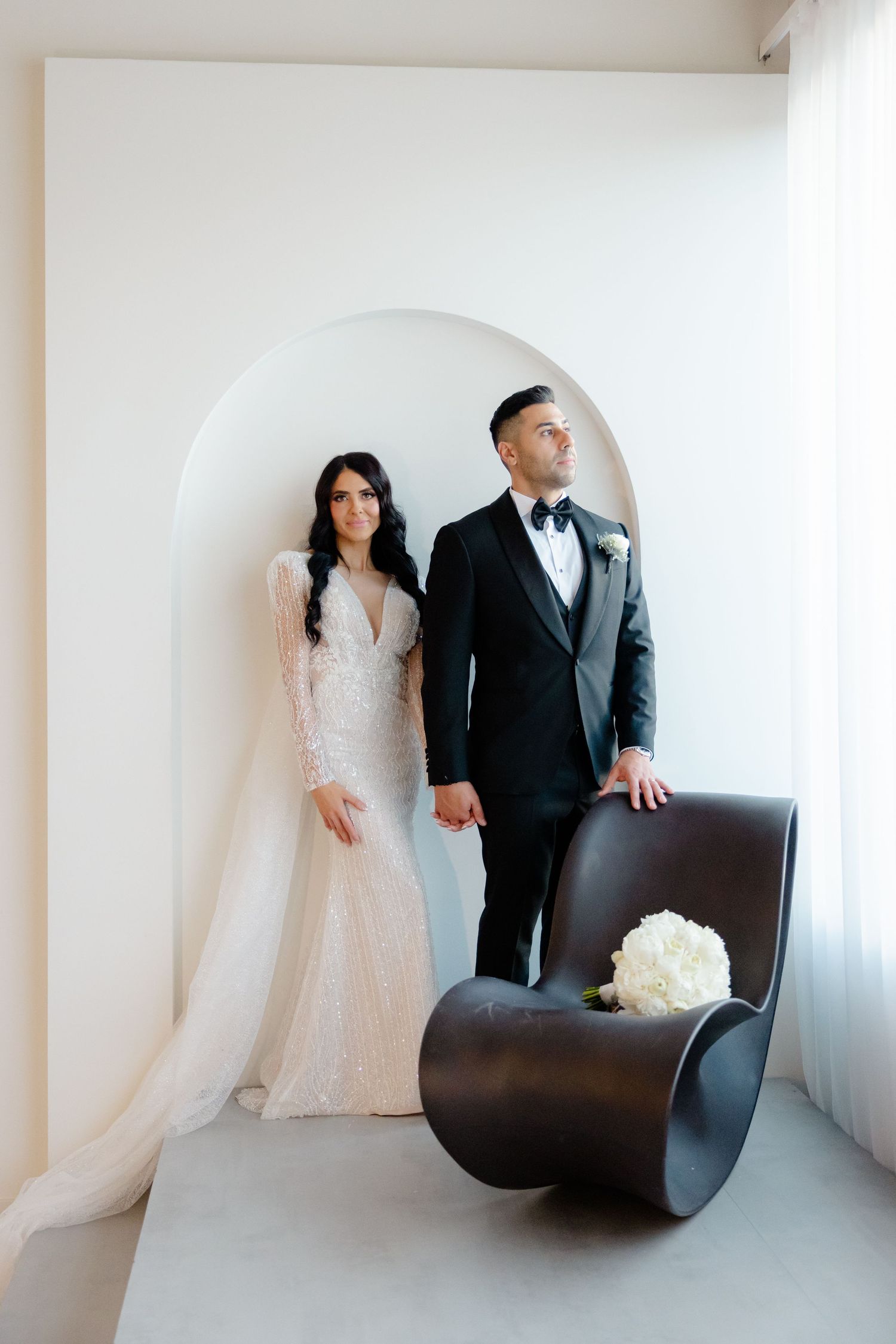 Wedding couple in formal attire standing in modern white setting with sculptural brown chair.