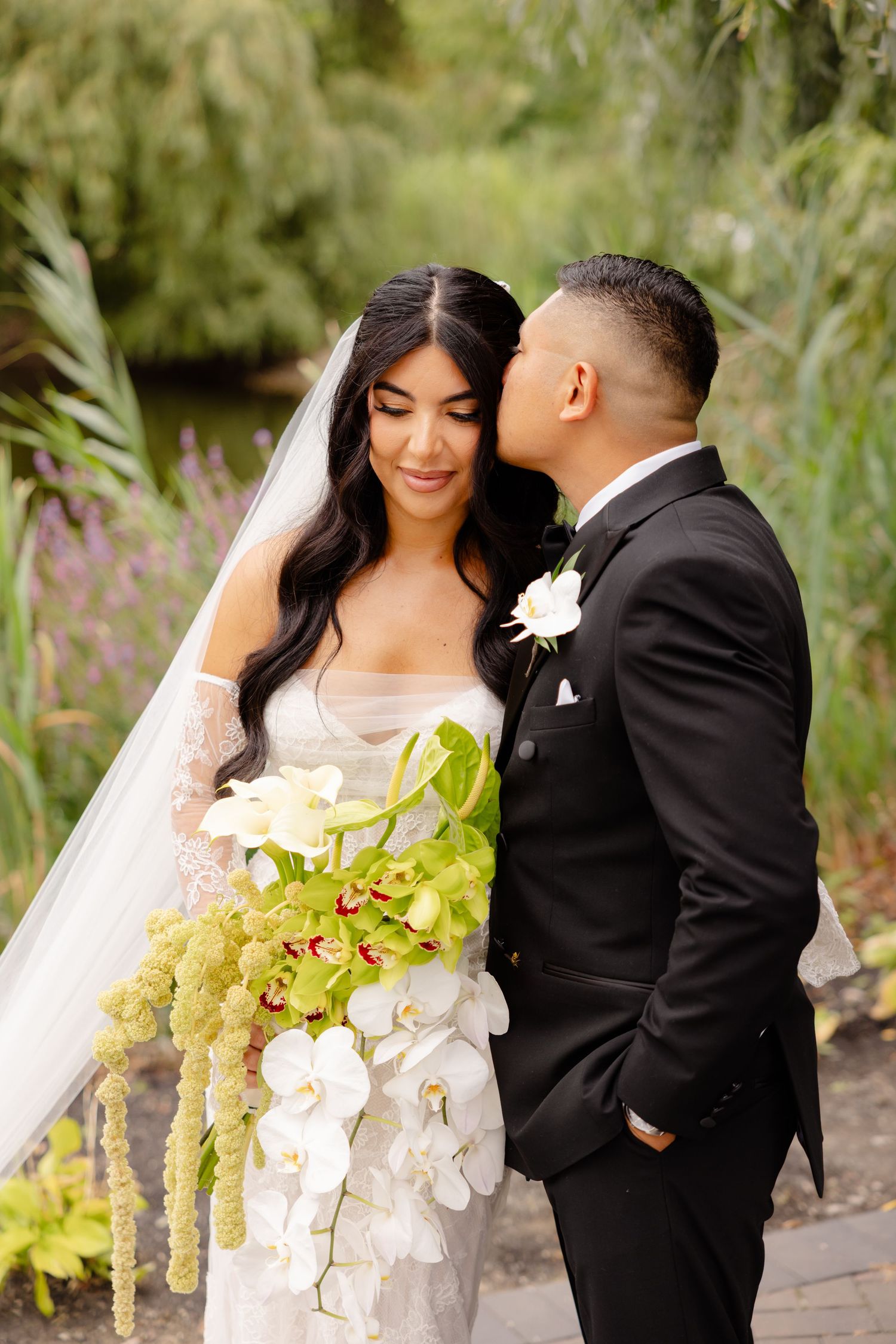 Bride in white lace gown with orchid bouquet and groom in black tuxedo share intimate moment in garden setting.
