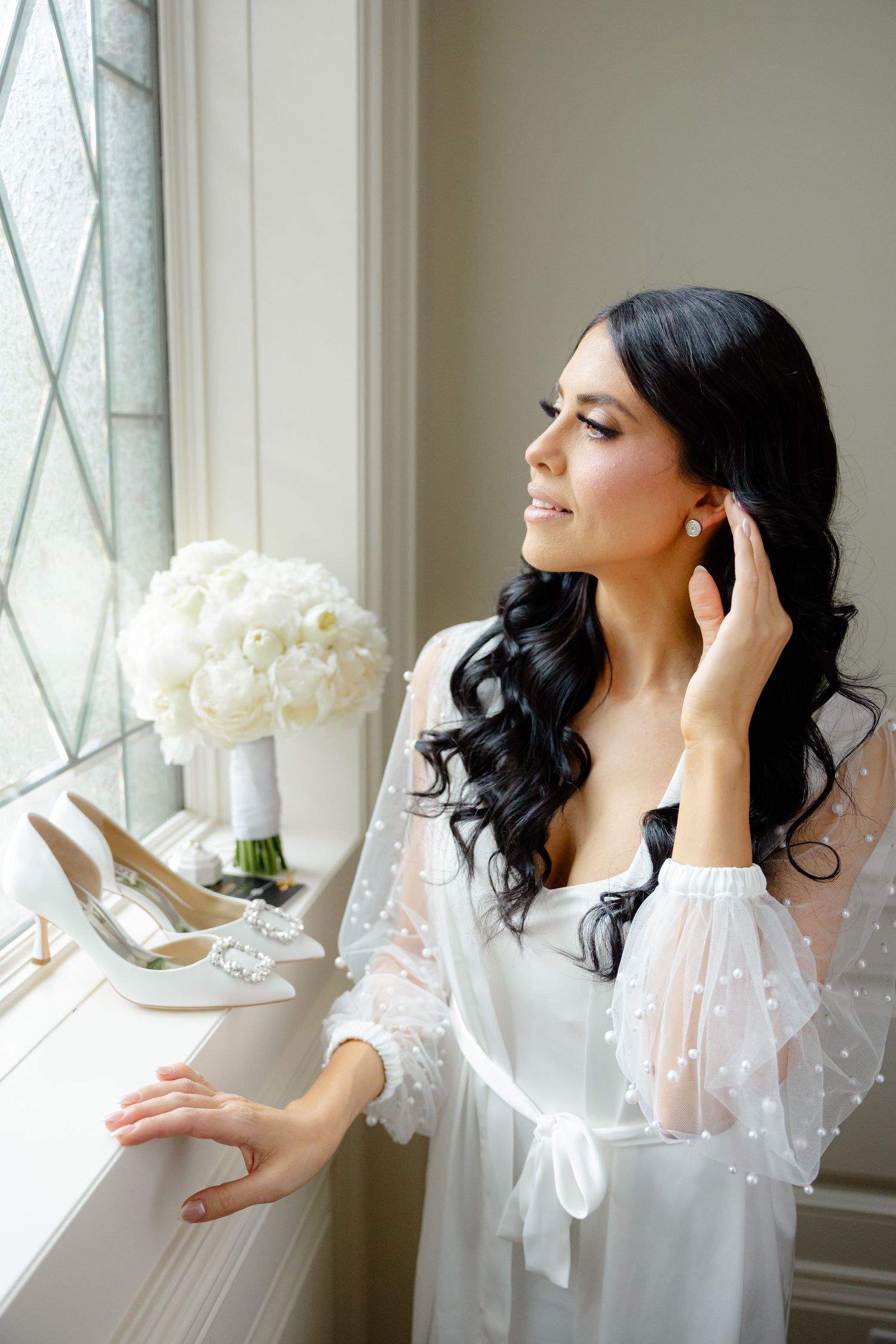 Bride preparing by window with white bouquet and wedding shoes on windowsill.