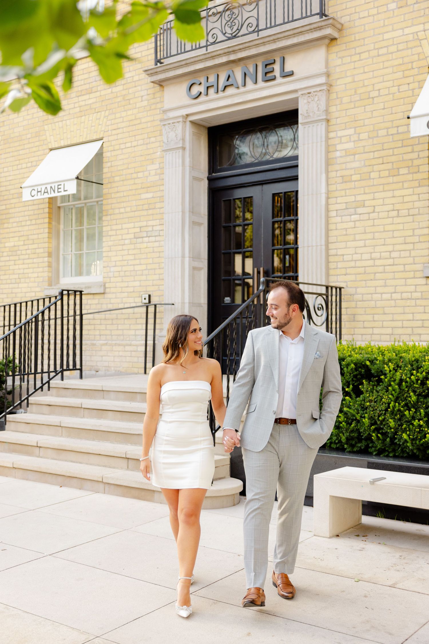 Couple holding hands while descending steps of upscale Chanel boutique on sunny day.